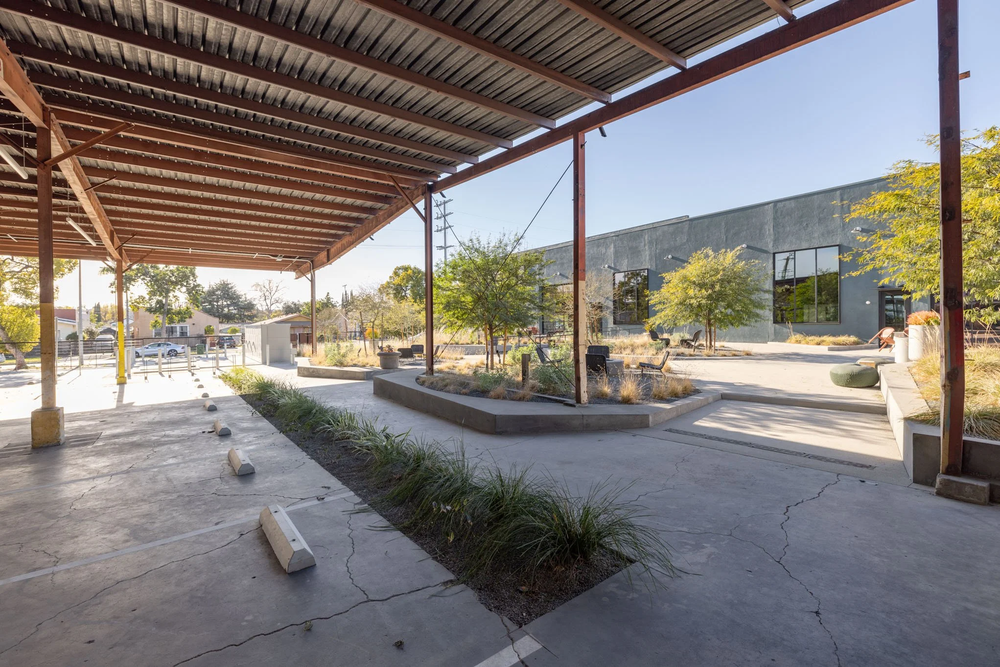 Empty parking lot with a shaded area, small trees, plants, benches, and a modern gray building in the background under a clear sky.