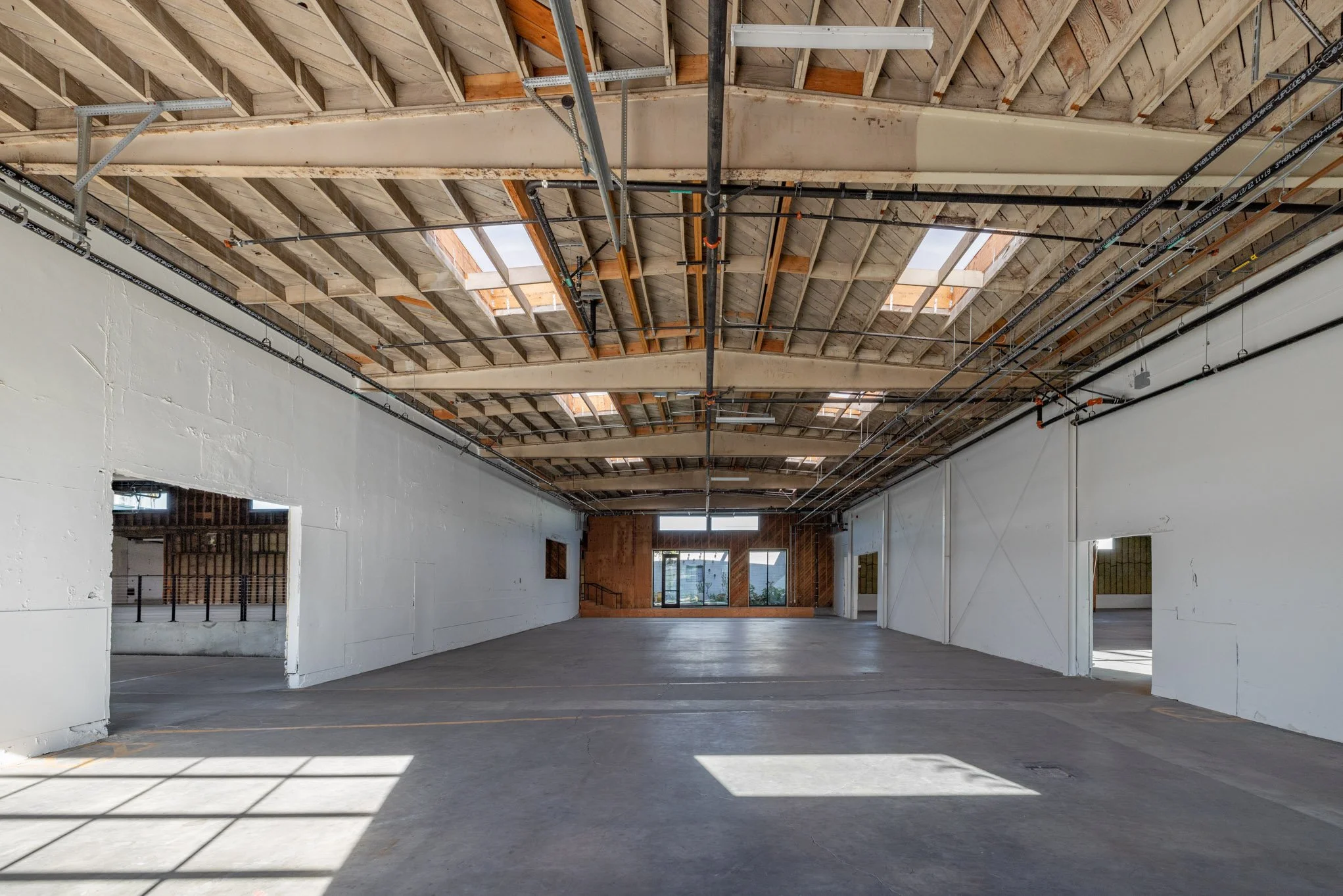 Empty interior space under construction with visible ceiling framing, skylights, and large windows, with white walls and concrete floor.