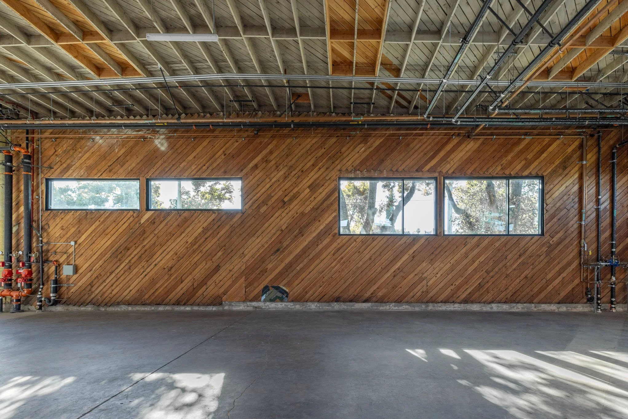 Empty industrial space with a wooden accent wall, three horizontal windows, and exposed ceiling pipes.