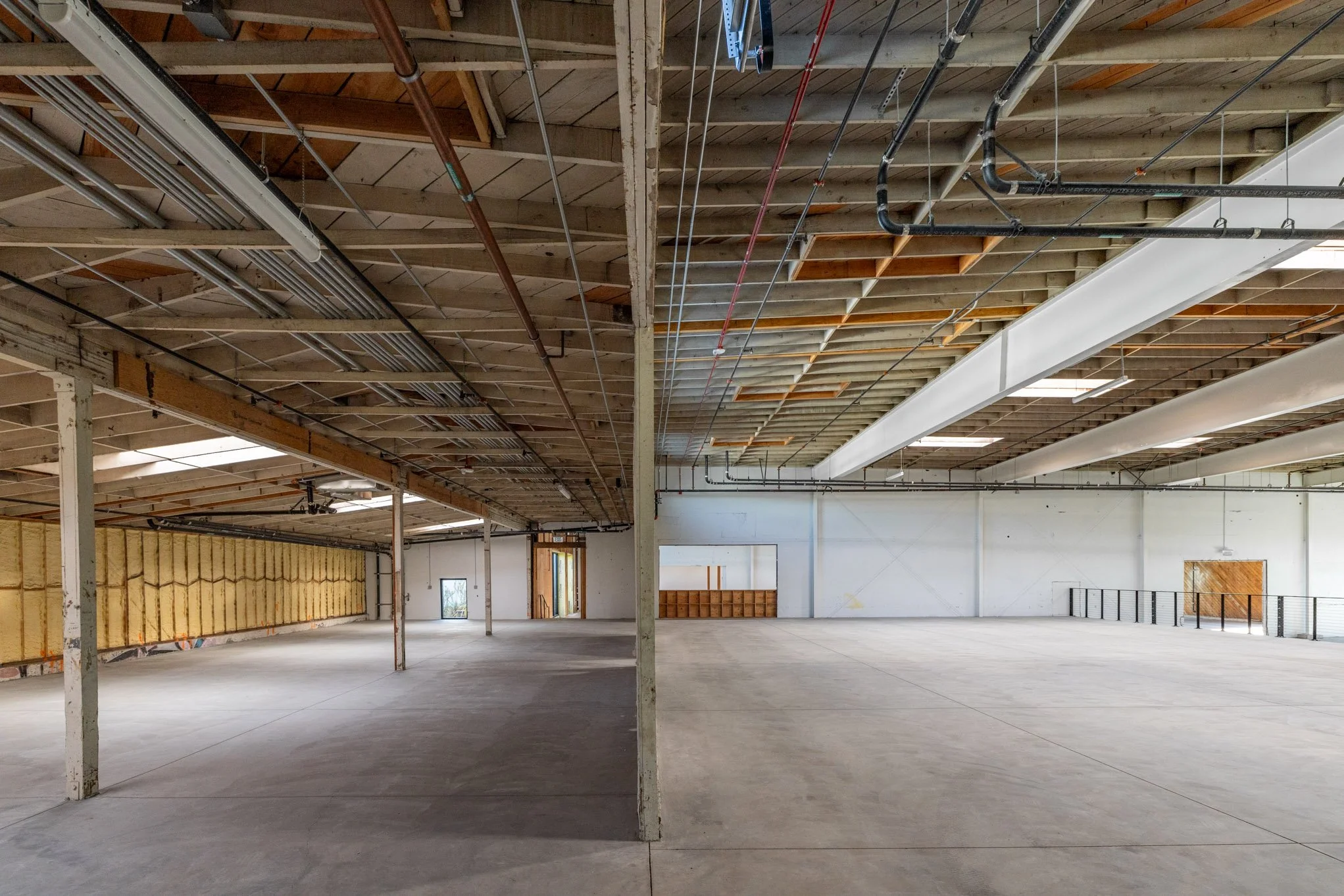 Interior of a large, empty warehouse with unfinished ceiling, exposed pipes and wires, and concrete floor, divided into two sections by a central column.
