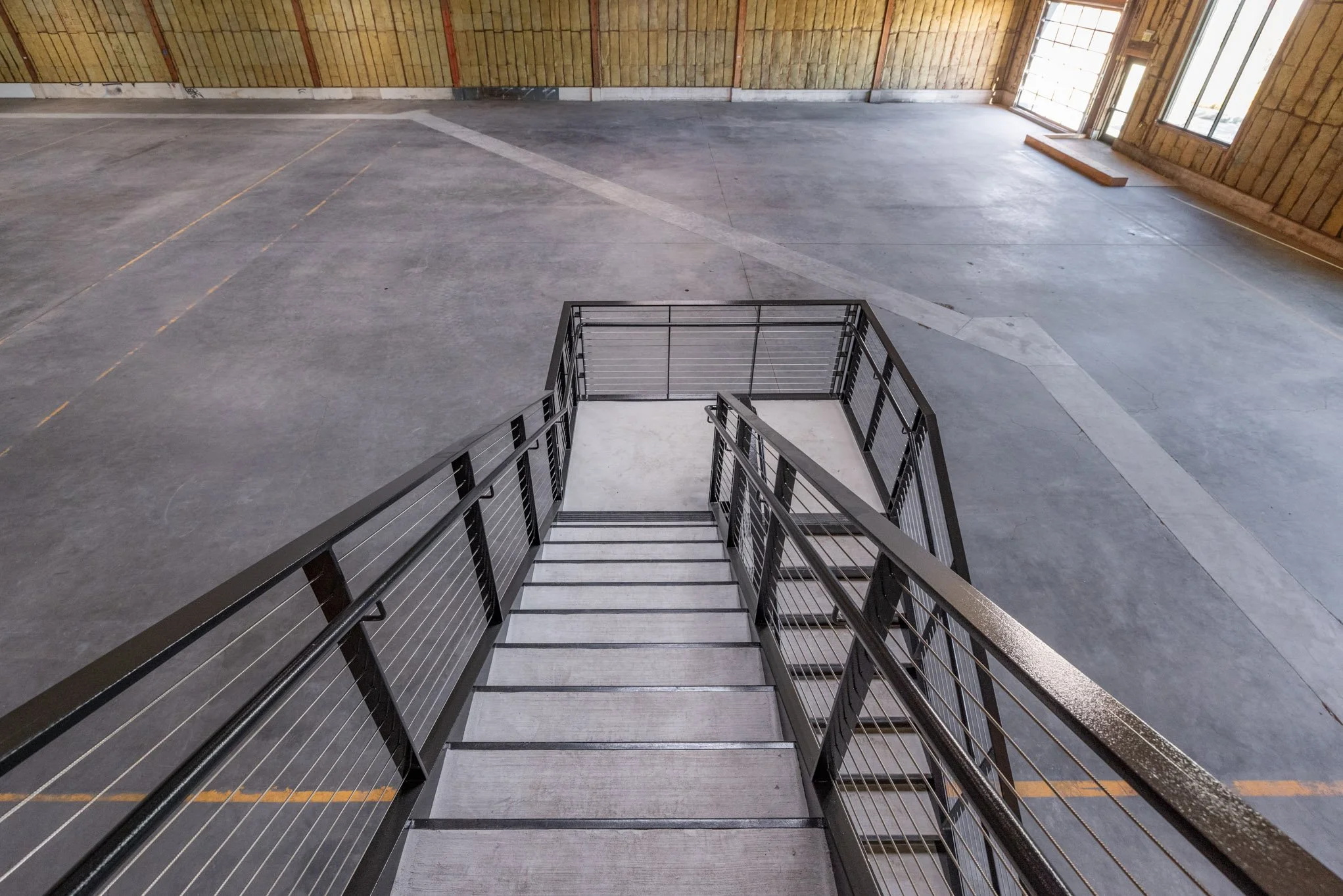Empty industrial parking garage with metal staircase and concrete floor and walls, sunlight coming through large window.