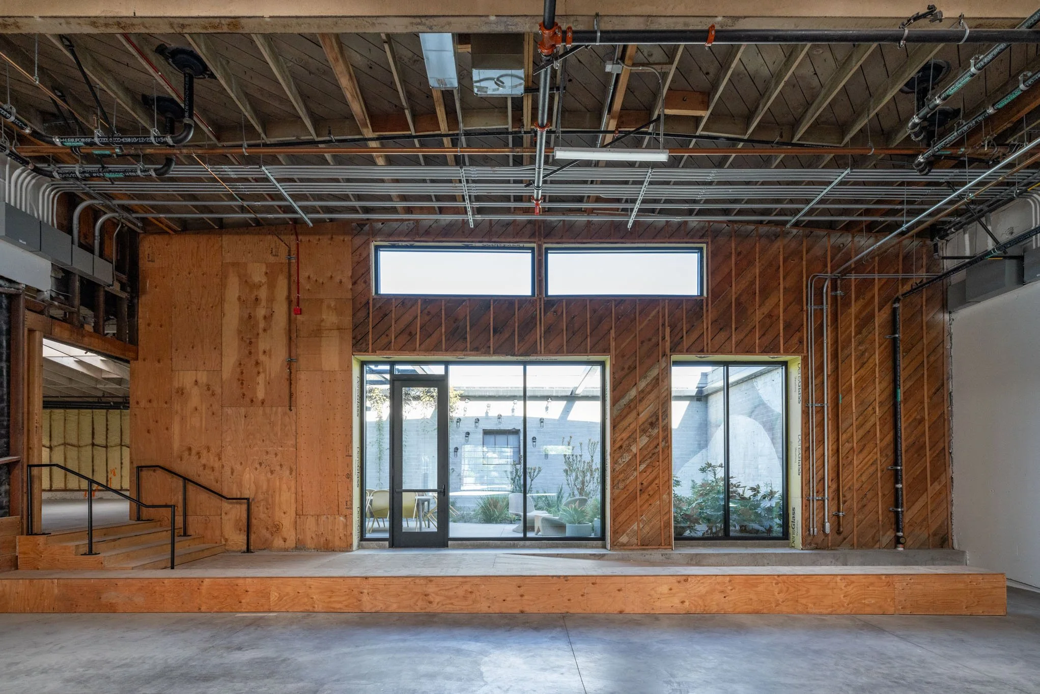 Interior of a building under construction with exposed wooden walls, ceiling framework, and electrical pipes, and a large glass door leading to an outdoor patio.