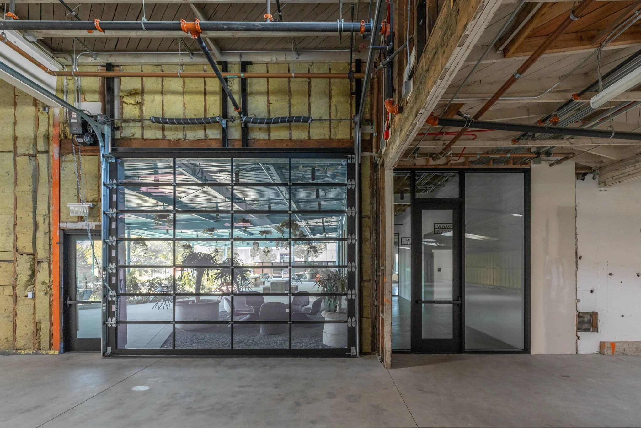 Interior of a building under construction with exposed framework, insulation, and piping. There are two glass doors, one with a grid-style frame, showing an area with plants and seating inside.