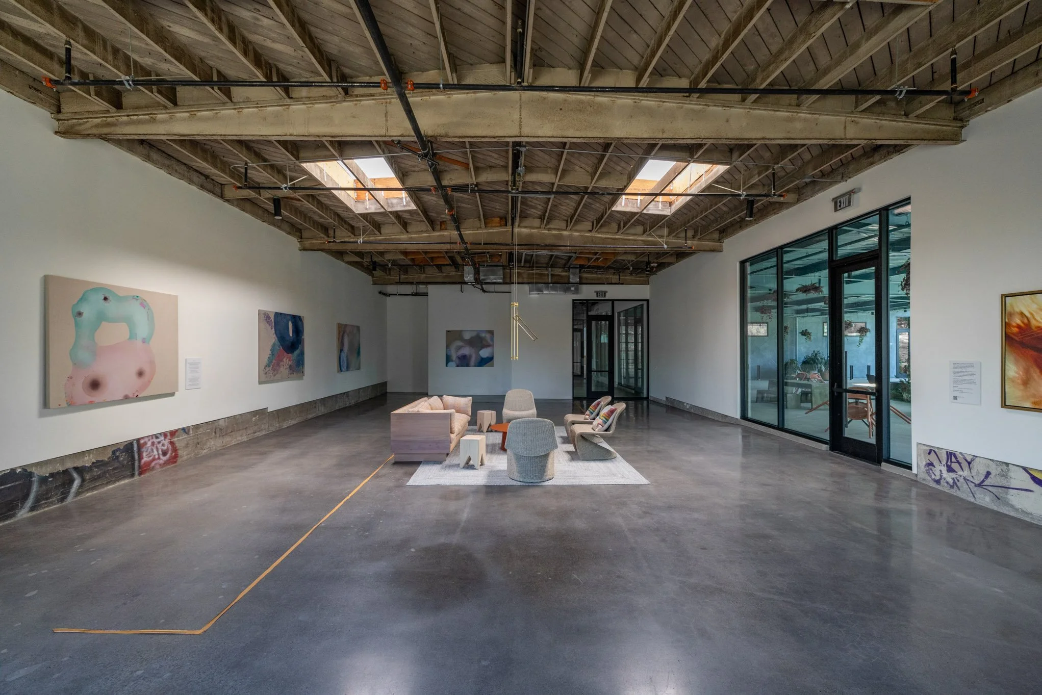 An art gallery with artwork on the walls, a seating area with chairs and a rug, and a large glass window/door on the right side.