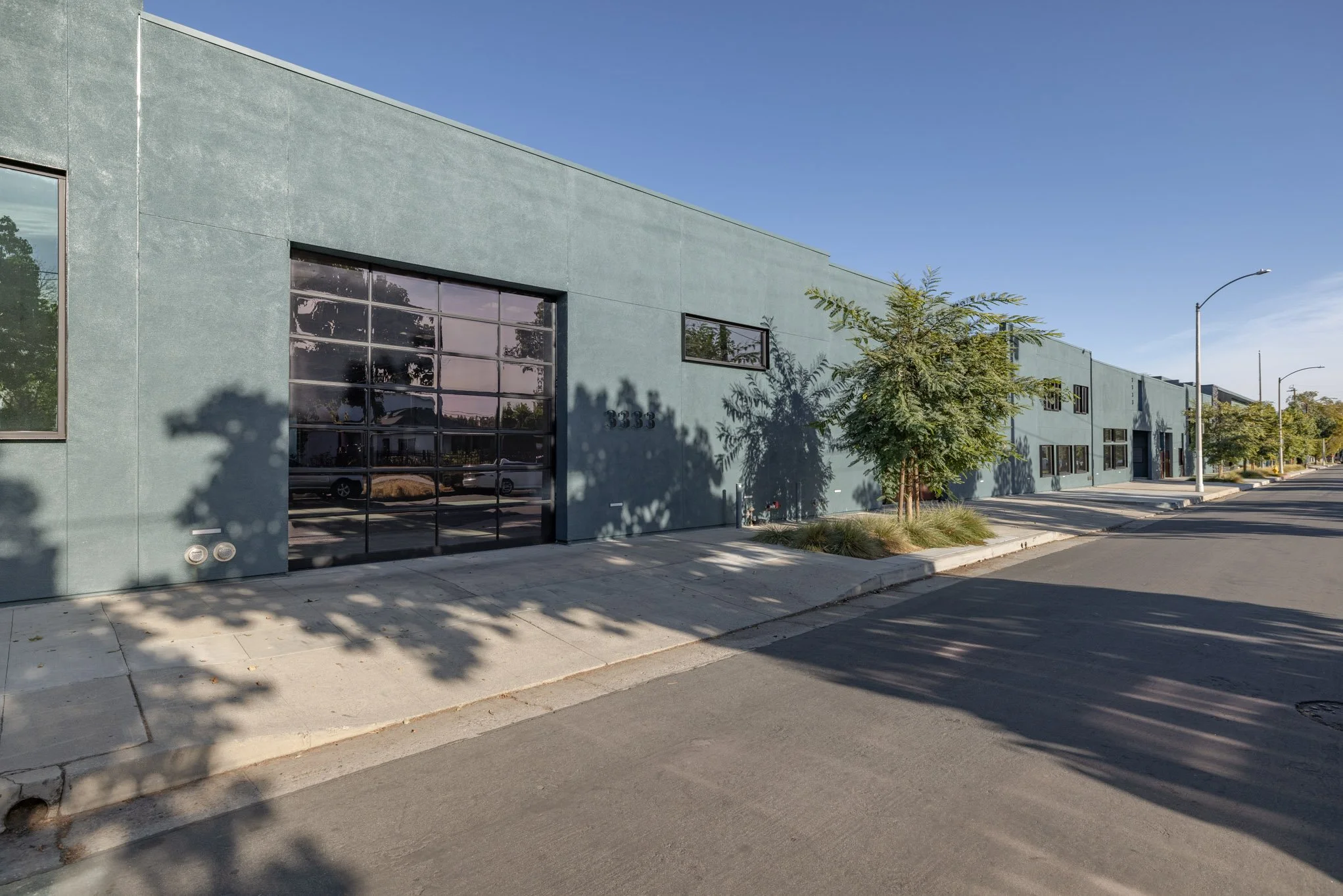 Empty commercial building with large glass garage door on a sunny day, trees planted along the sidewalk, and streetlights under a clear blue sky.