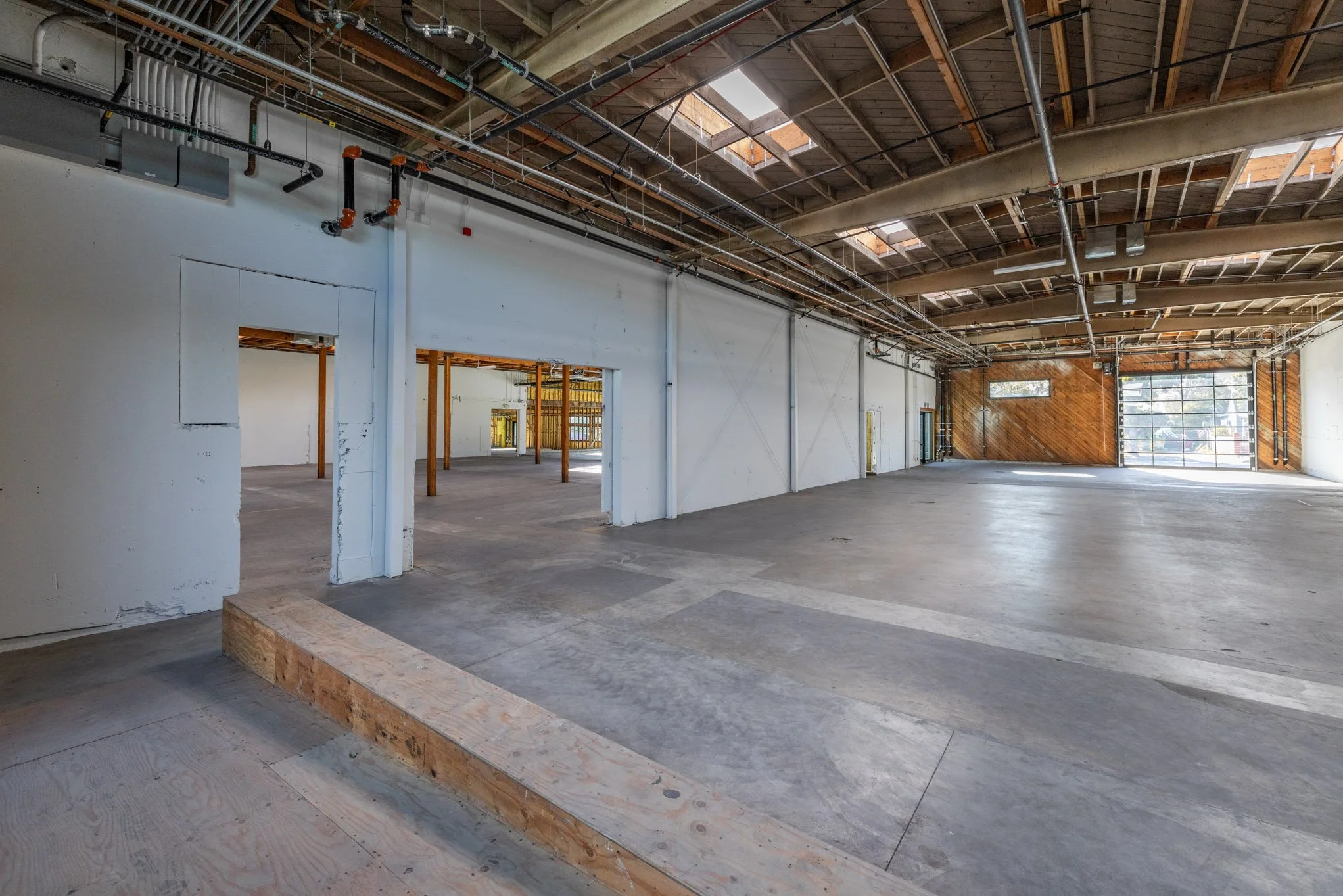 Empty interior of a commercial space with unfinished features, high ceilings with exposed beams, skylights, and large garage-style door, with some wooden and concrete elements.