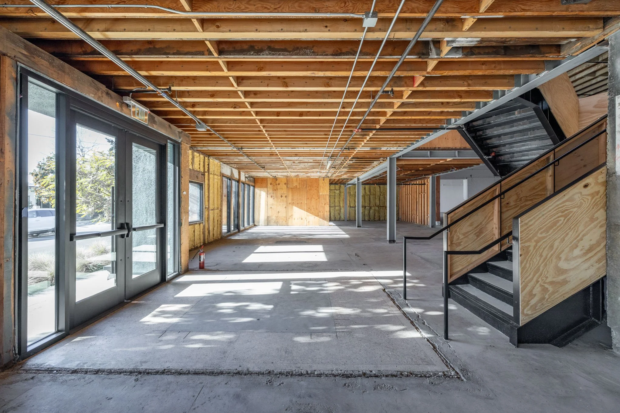 Interior view of a building under construction with exposed wooden beams, large glass windows and doors, a staircase with black railing and wood panels, and insulation on the walls.