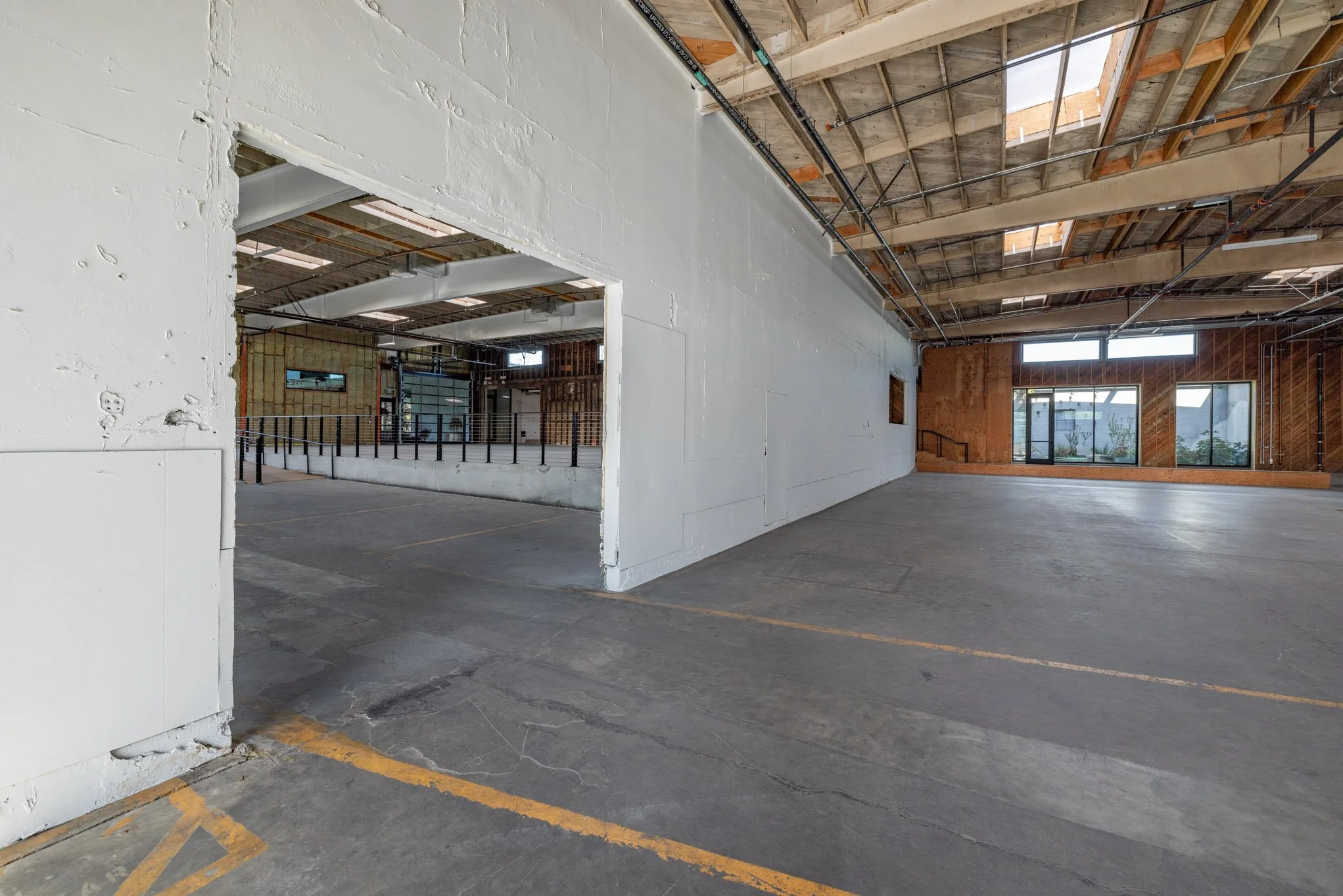 Empty industrial or commercial warehouse space with concrete floors, unfinished ceilings, and large windows allowing natural light.