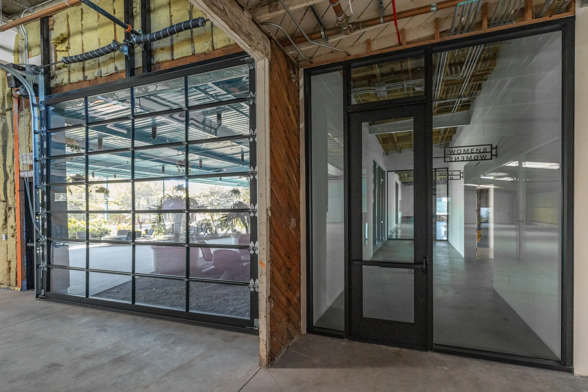 Image shows a partially constructed indoor space with a garage door on the left and a glass door leading to a hallway or room on the right. The garage door is made of glass panels with an industrial design, and the hallway has signs indicating it lea