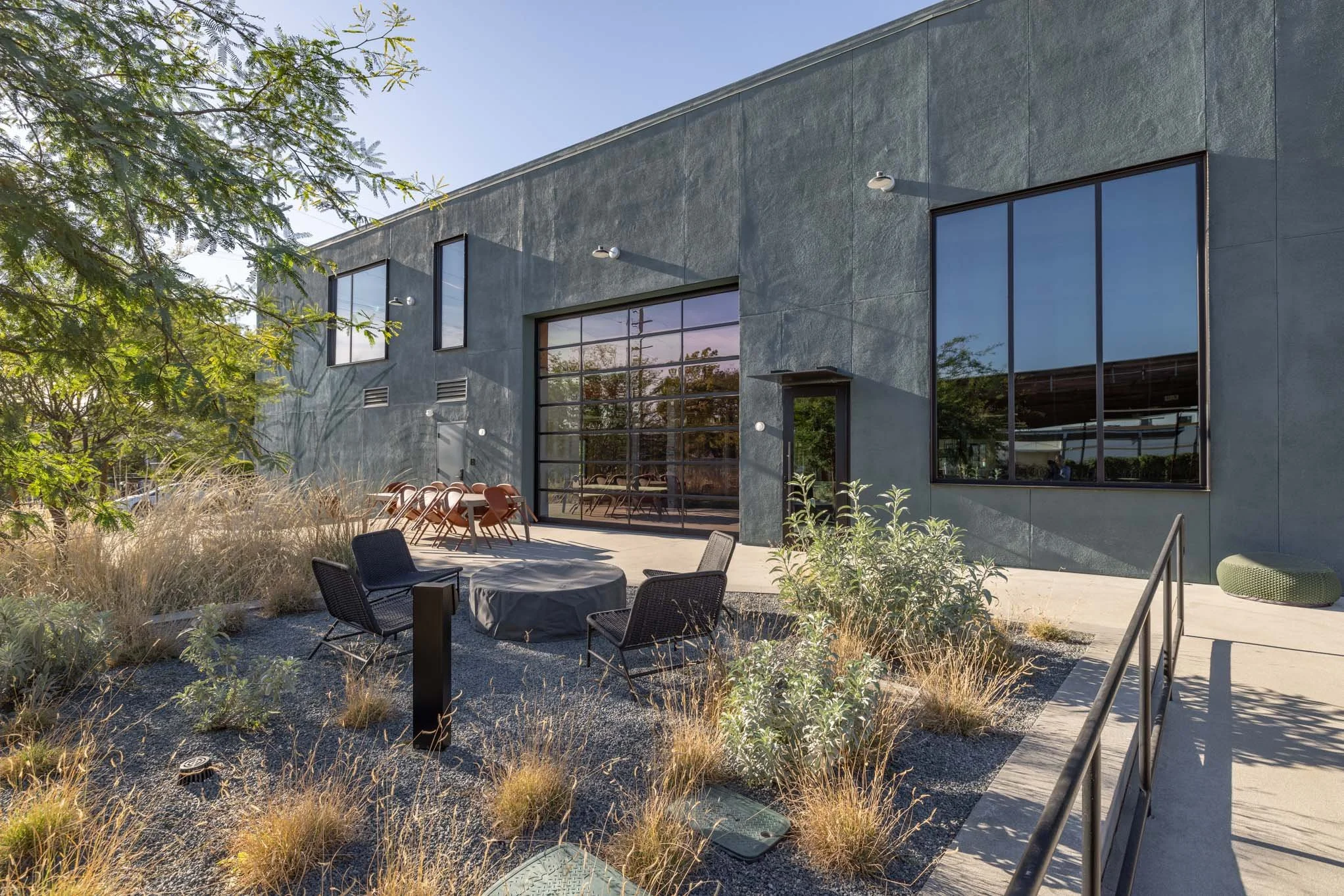 Modern industrial building exterior with large glass windows and patio seating, surrounded by desert landscaping and a clear blue sky.