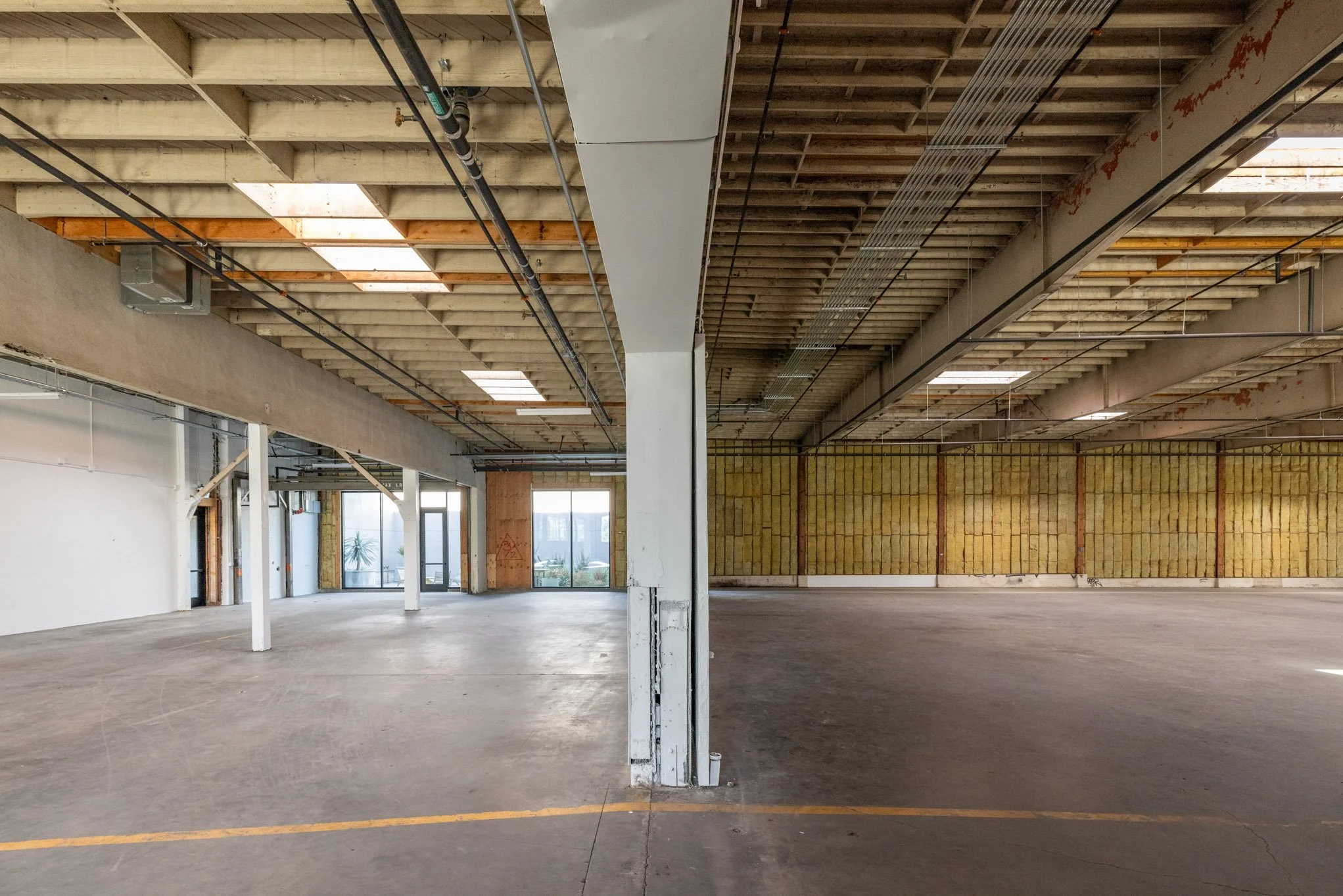Empty, partially renovated industrial or commercial space with exposed ceiling beams, plywood, and insulation, divided by a central support column.