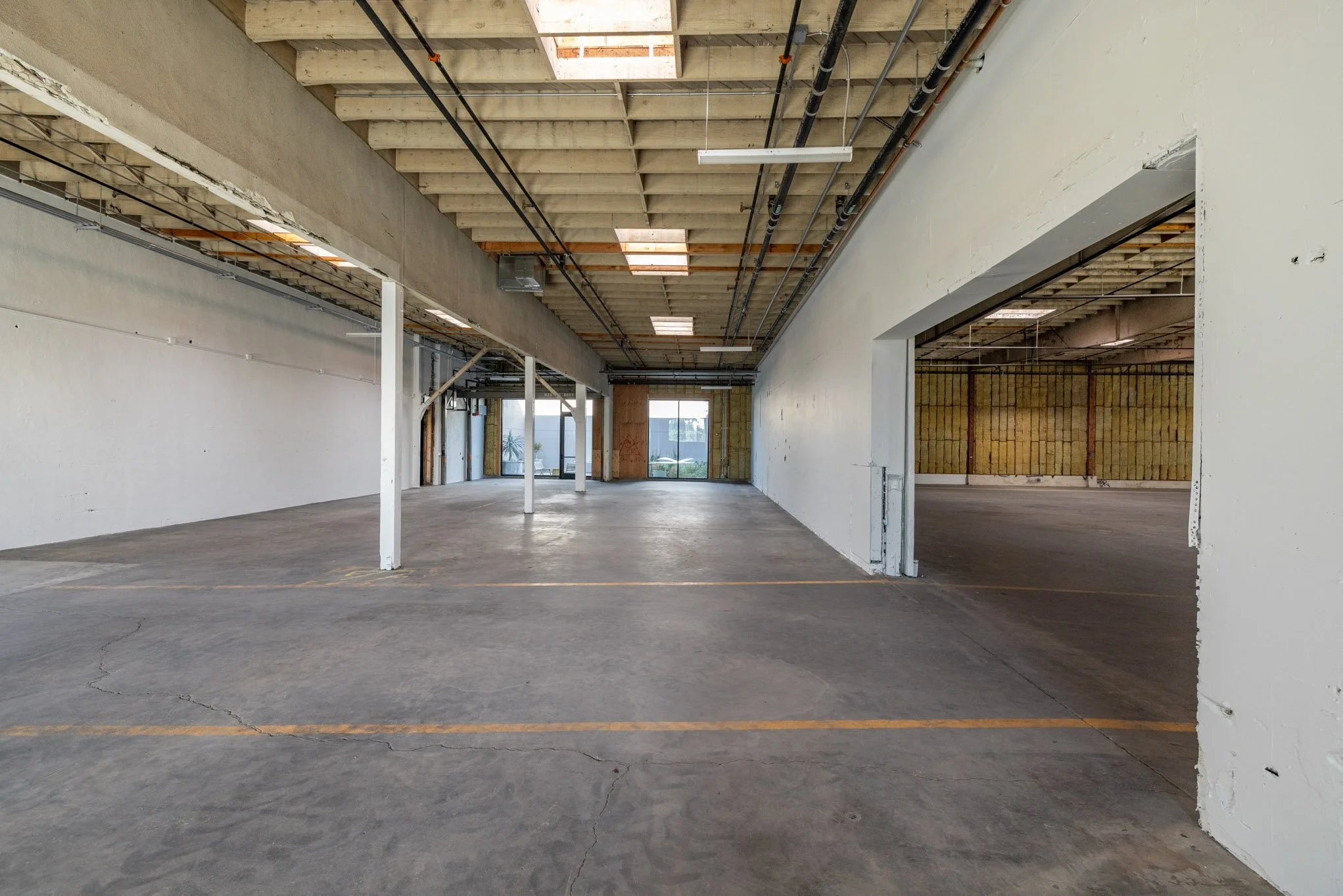 Empty unfinished commercial or industrial space with concrete floors, white painted walls, exposed ceiling with roofing panels, electrical wiring, and support beams.