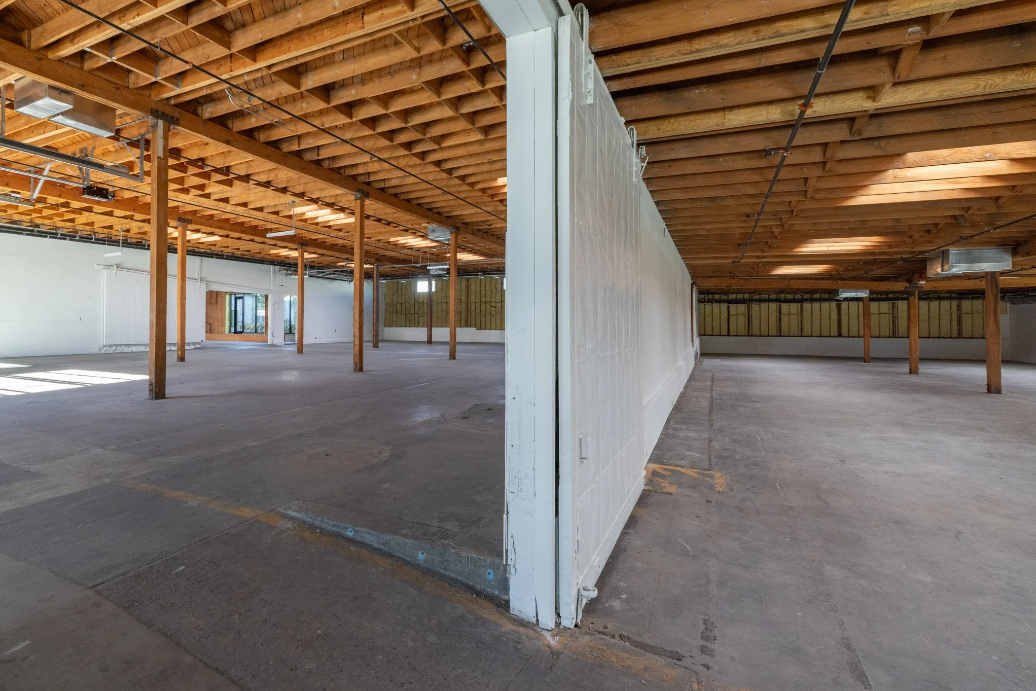 Interior of a building under construction with exposed wooden ceiling beams, support columns, and unfinished concrete floors, separated by a partial wall.