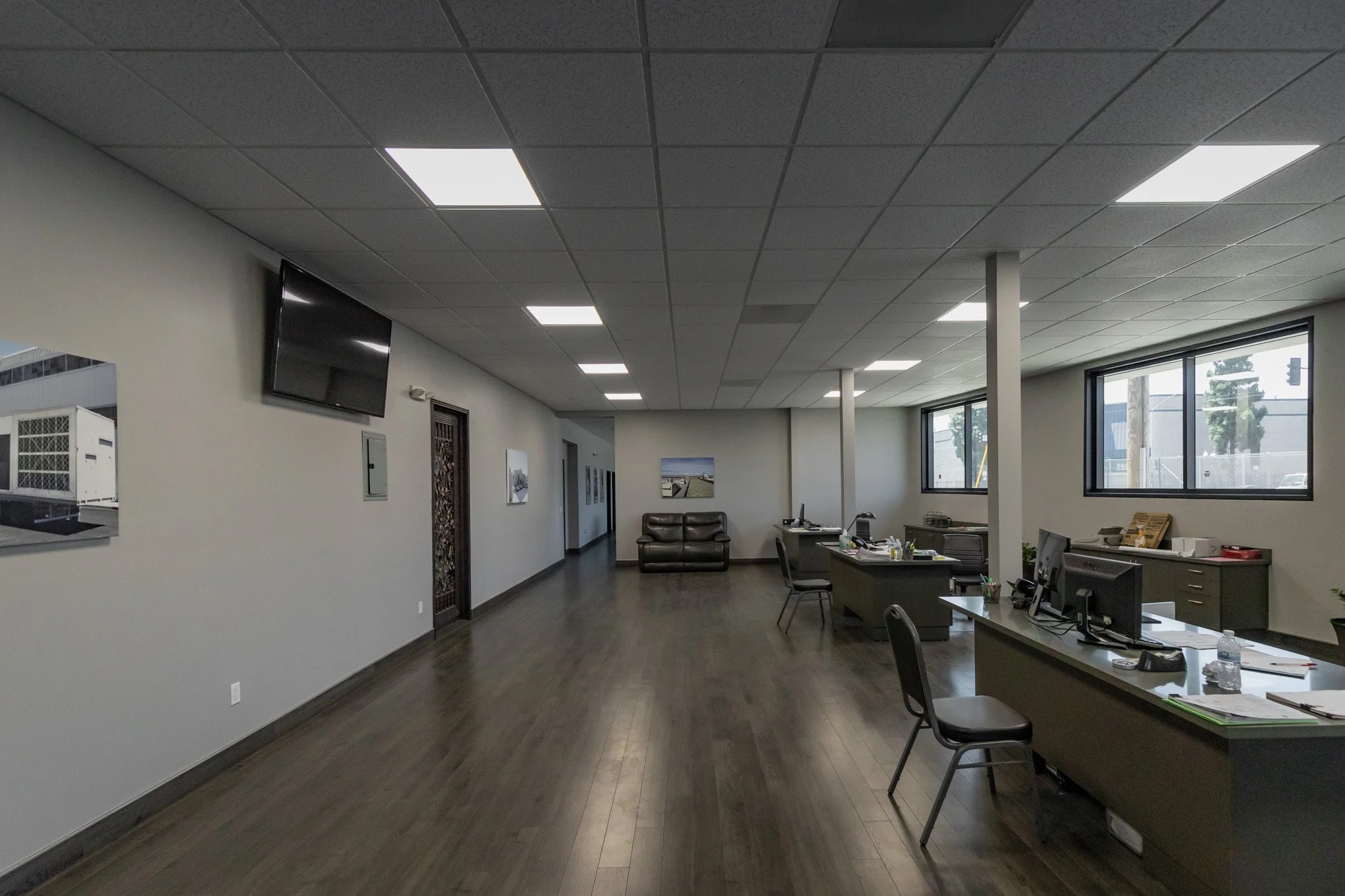 Empty office with desks, chairs, computers, large windows, and wood flooring.