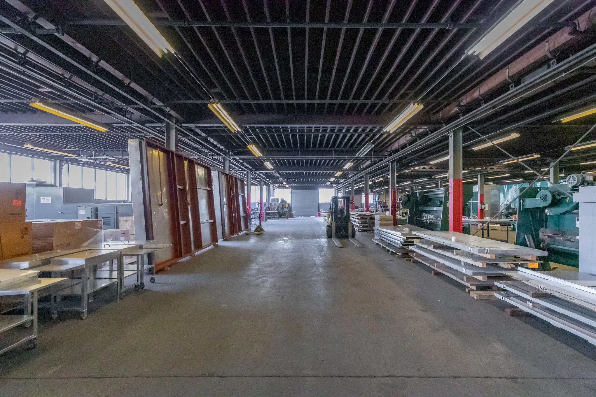 Interior of a warehouse or workshop with metal shelving, construction materials, and machinery, illuminated by fluorescent lights.