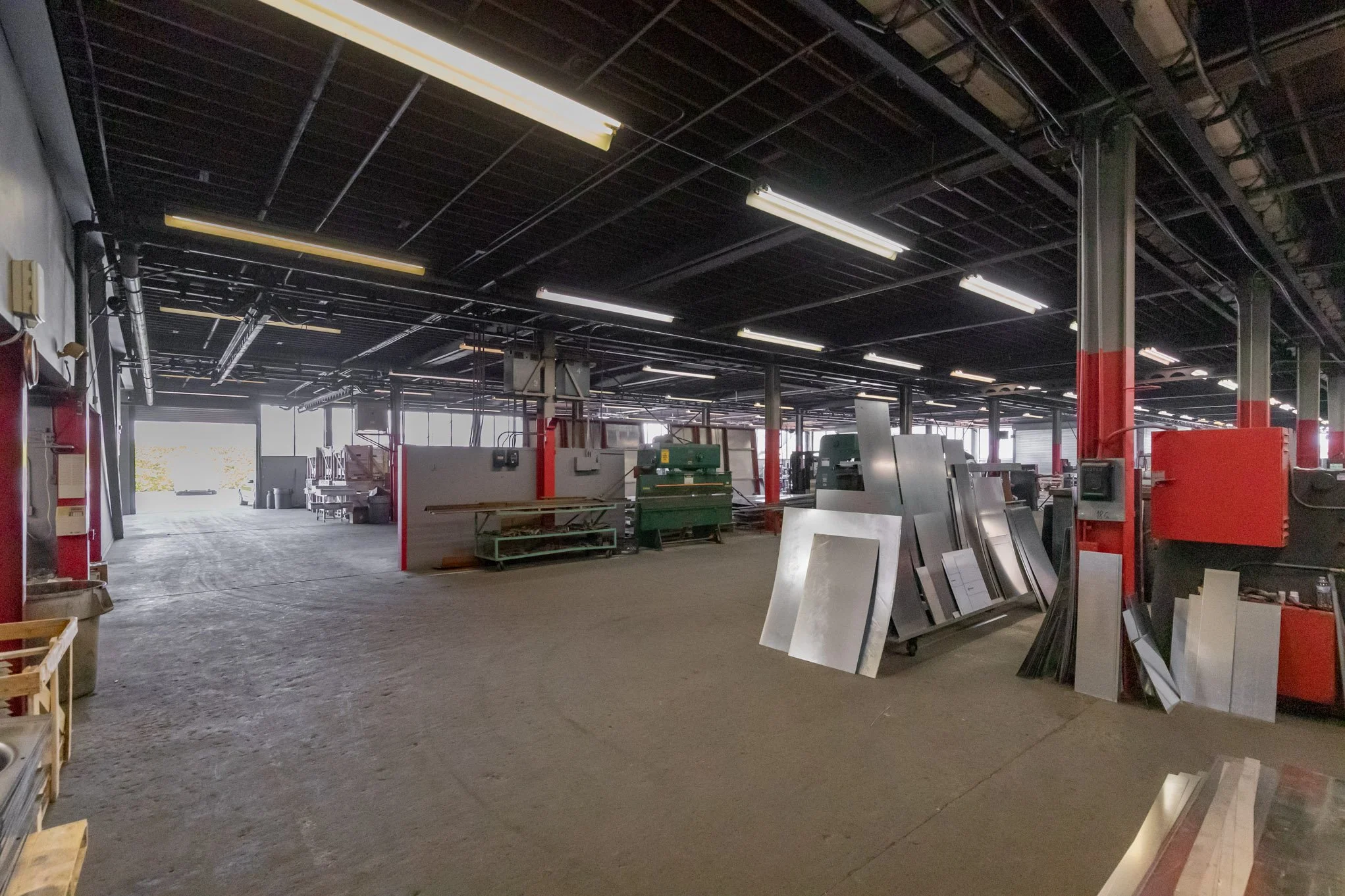 Industrial warehouse with metal sheets, tools, and equipment. Open entrance shown at the back, with sunlight streaming in.