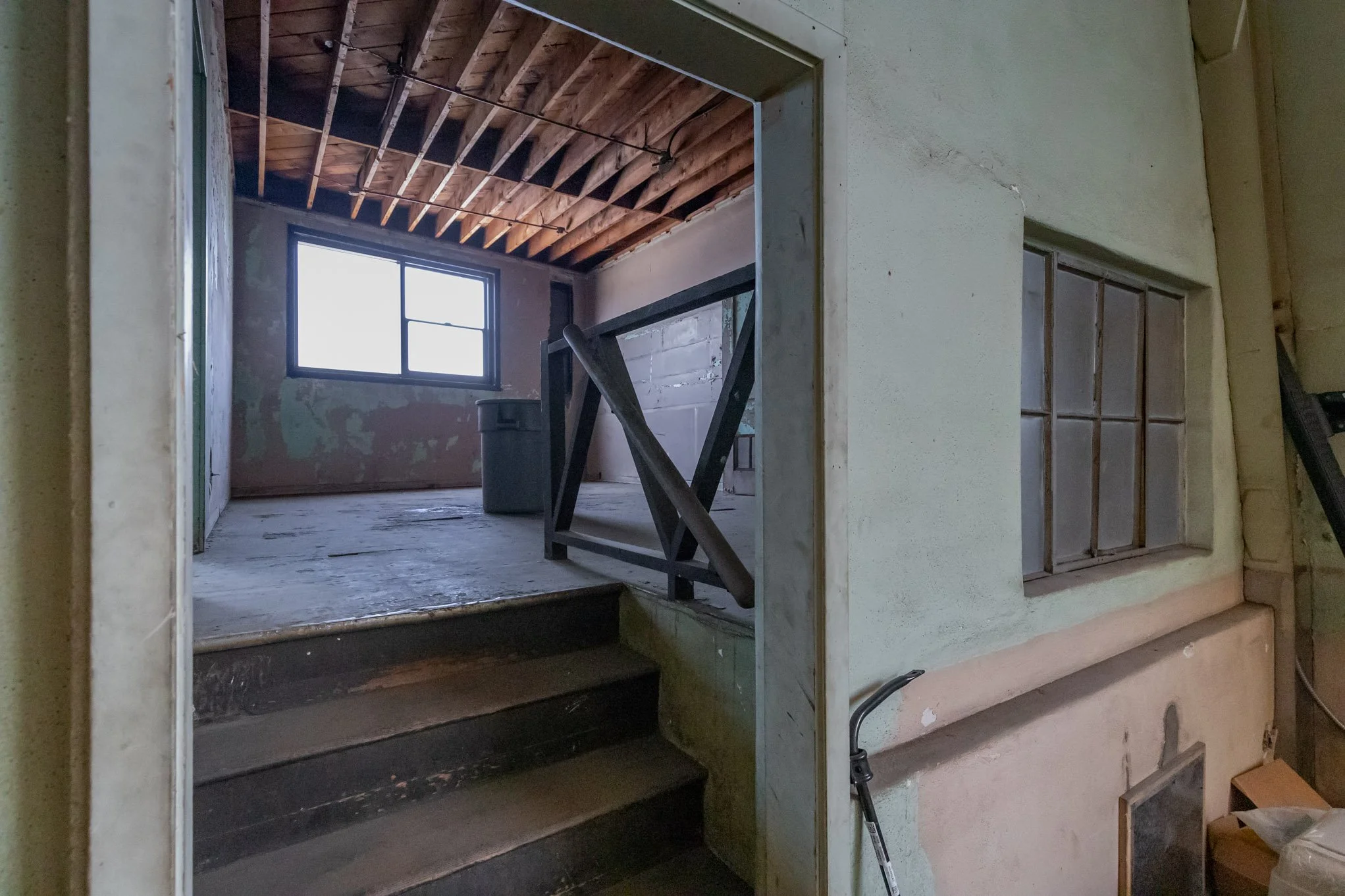 Interior of an unfinished or abandoned room with an open doorway, showing stairs leading up to a small room with a window, worn walls, and exposed wooden ceiling beams.