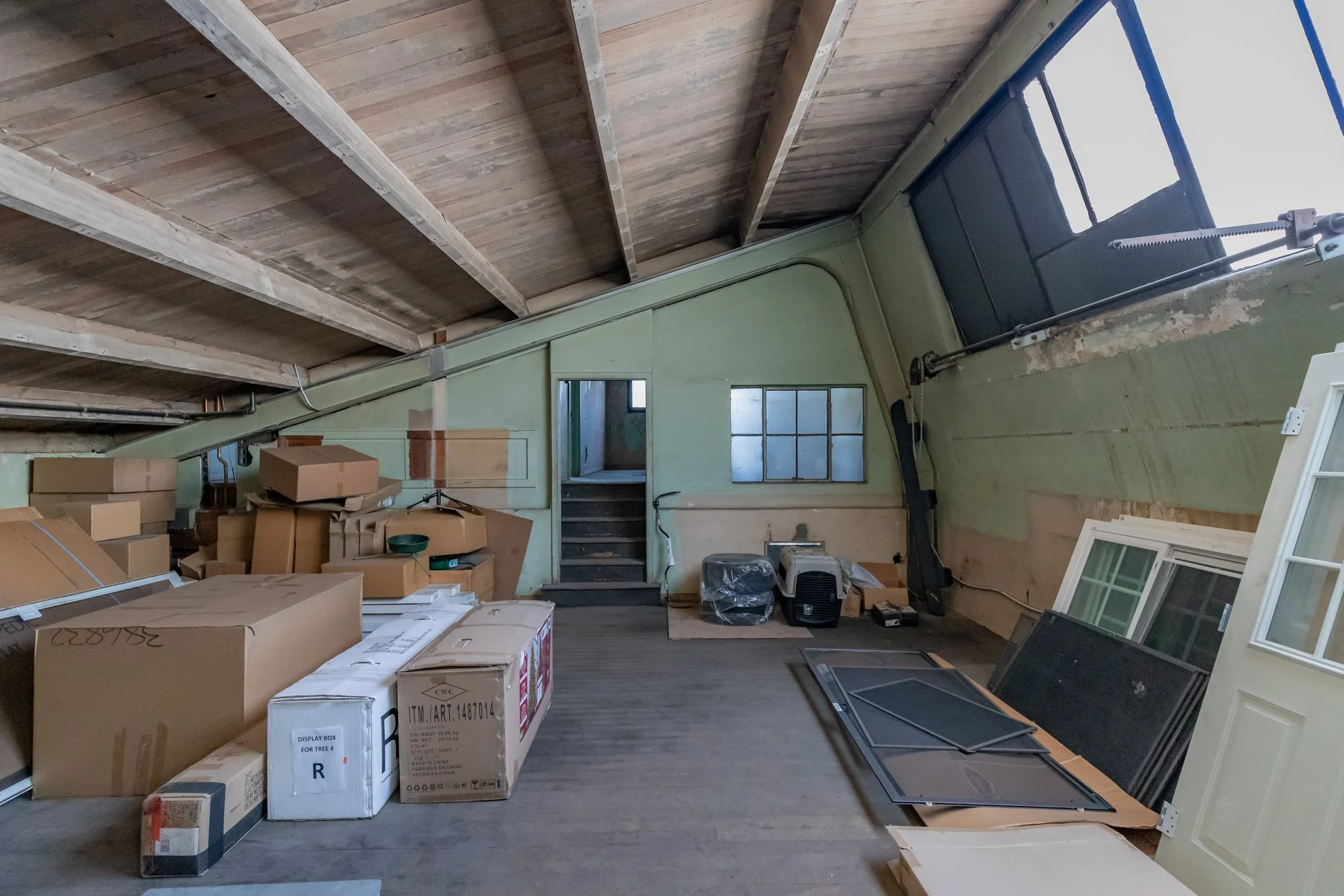 An attic room filled with cardboard boxes, window frames, and other miscellaneous items, with a sloped wooden ceiling and a small step leading upstairs.