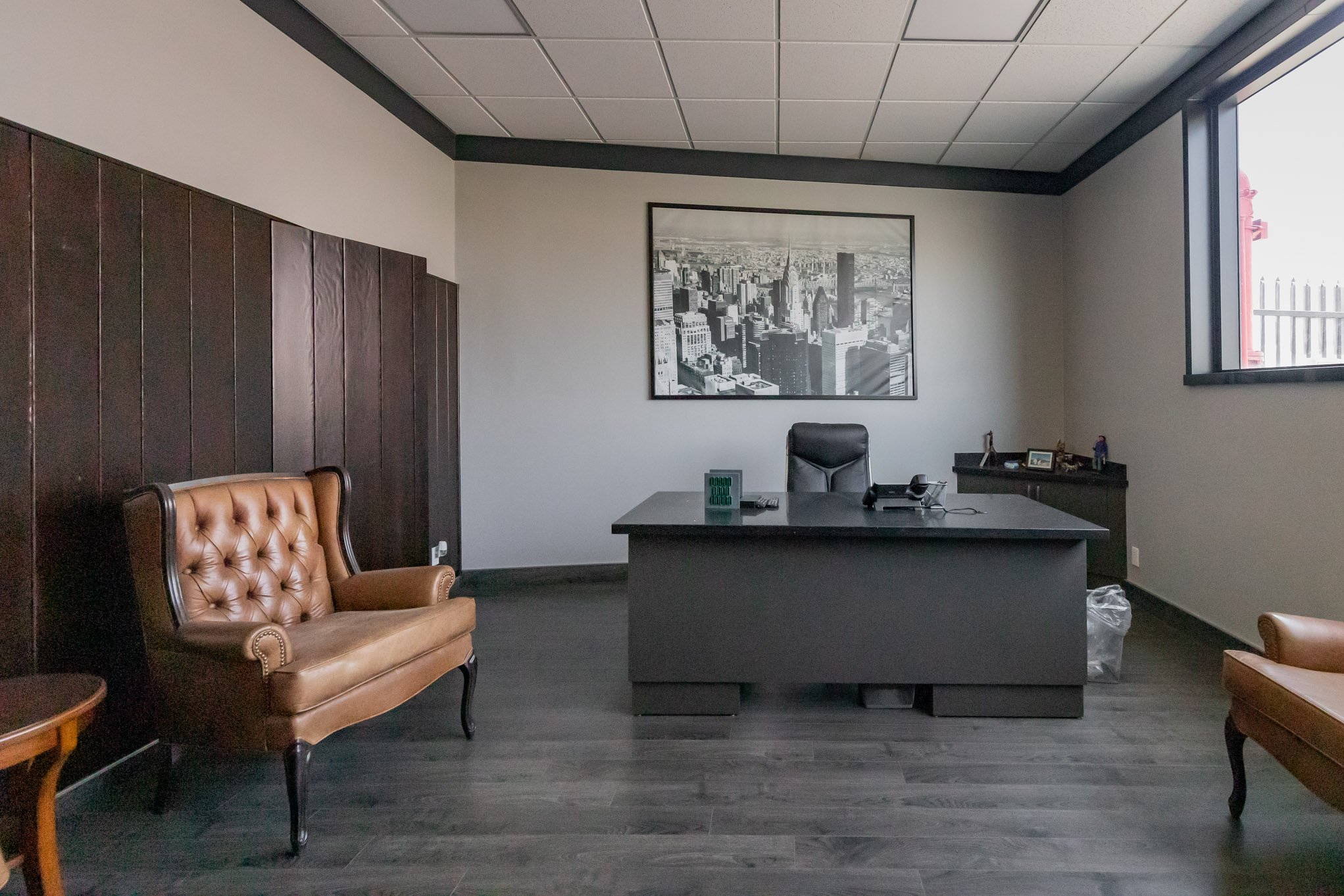 Empty modern office with a black desk, leather chairs, framed cityscape on the wall, window with a view outside, and dark wood paneling.