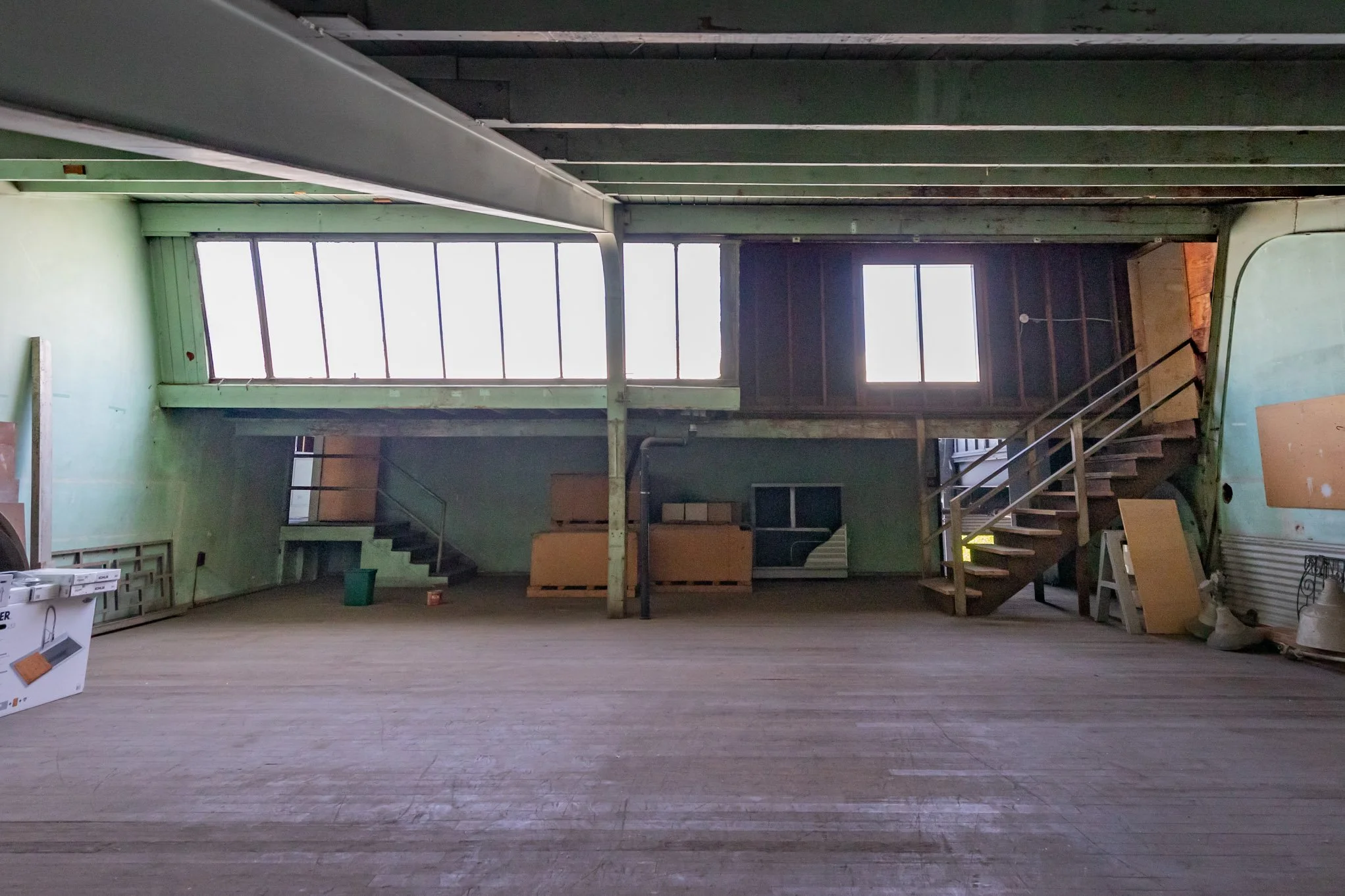 Interior of an unfinished, empty building with green walls, wooden floors, stairs on both sides leading to a second level, large windows letting in natural light, and construction materials scattered around.