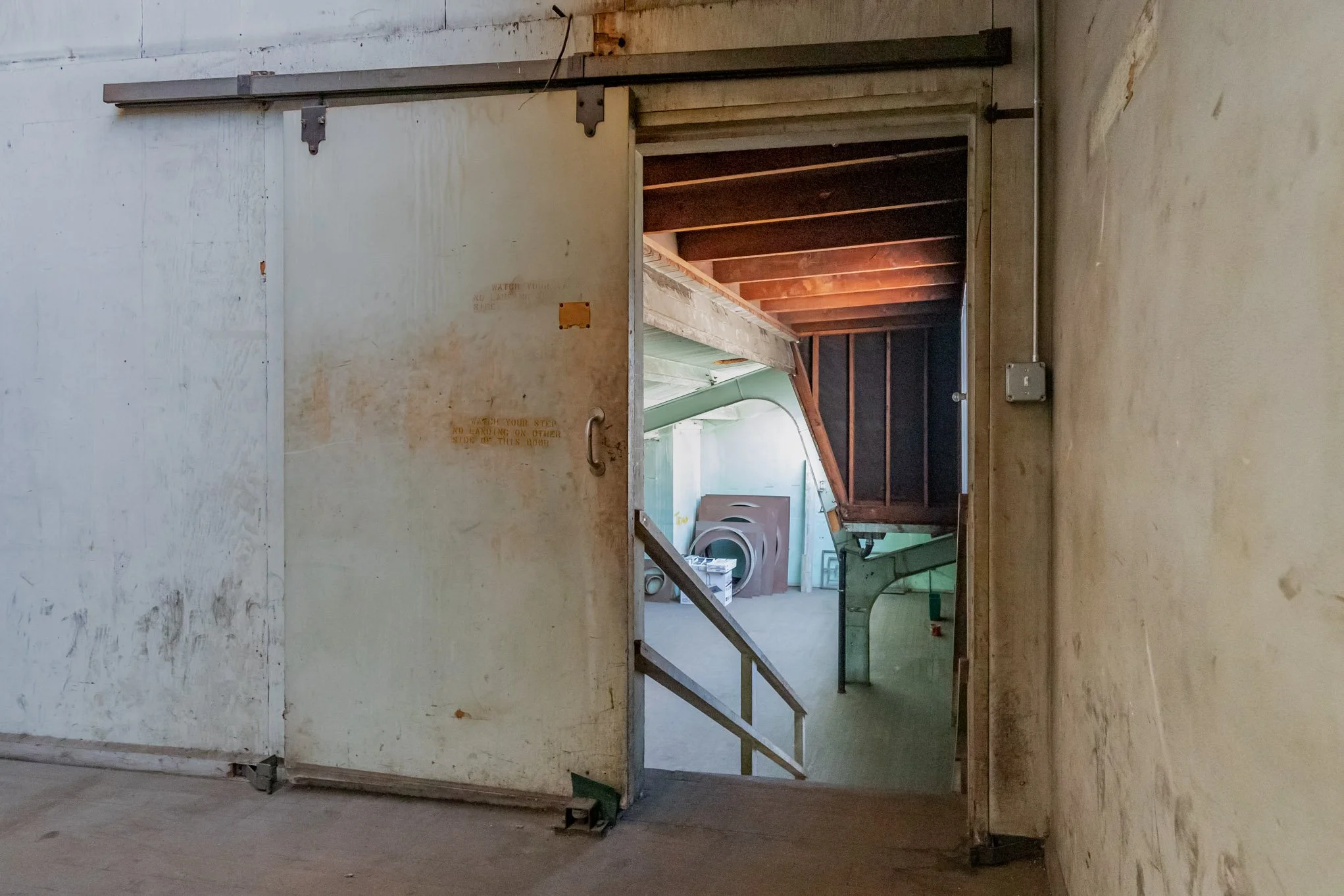 View through a partially open sliding door into a storage or laundry room with stacked washers and dryers, visible pipework, and unfinished wooden ceiling beams.