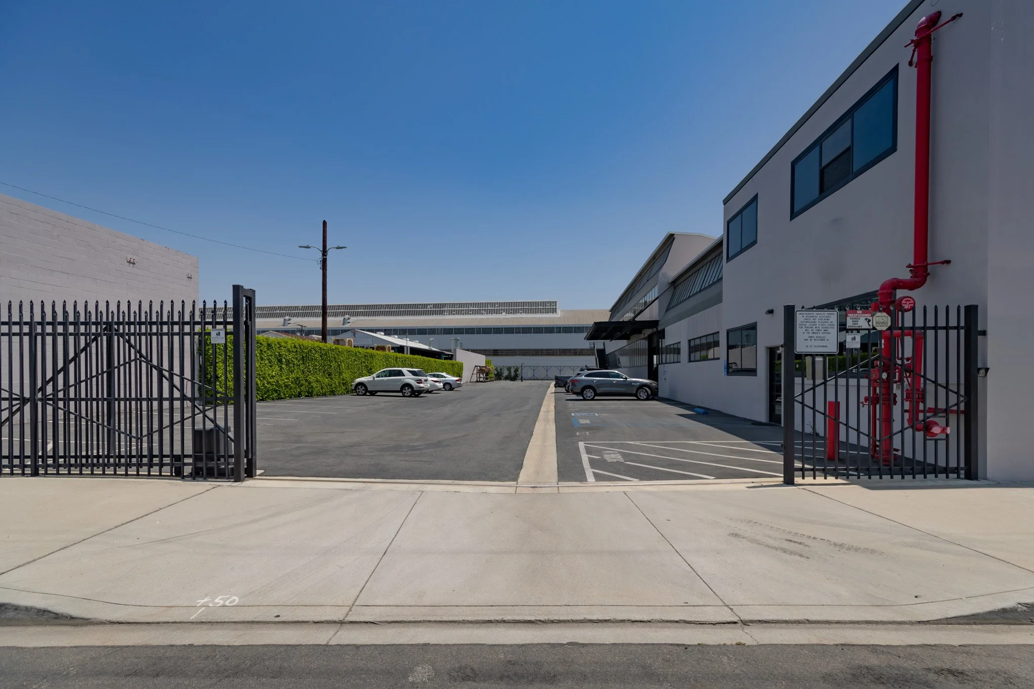 Empty parking lot next to a white industrial building, with a black fence and gate in the foreground, clear blue sky overhead.