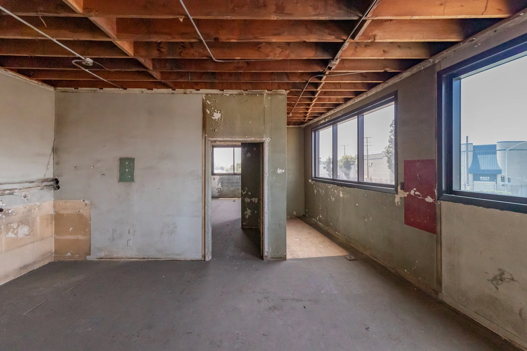An unfinished interior room with exposed wooden ceiling beams, traced electrical wiring, and partially painted and damaged walls. Large windows on the right side let in natural light, revealing adjacent buildings and street views.