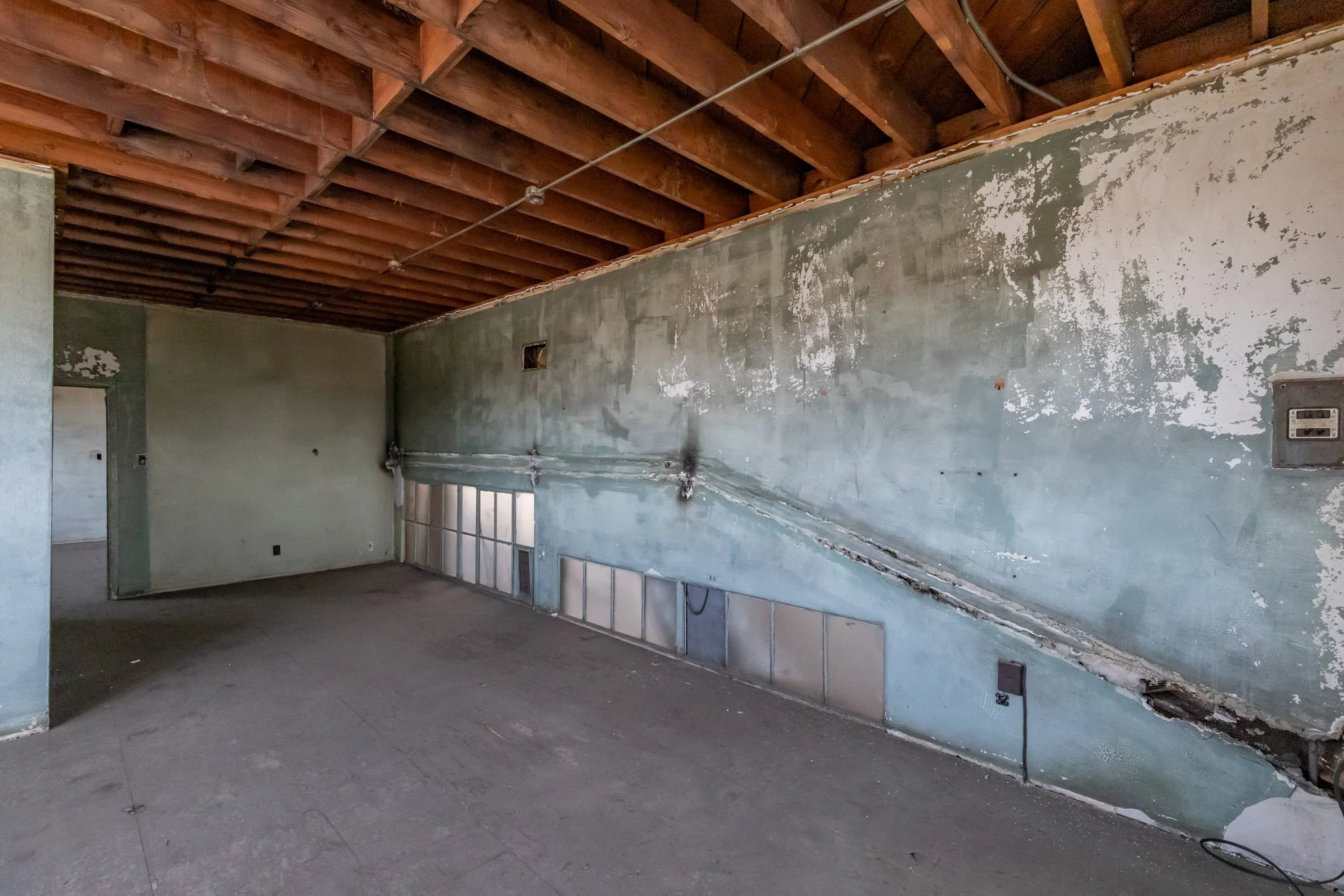 Empty room with worn green walls, exposed wooden ceiling, and boarded-up lower windows.