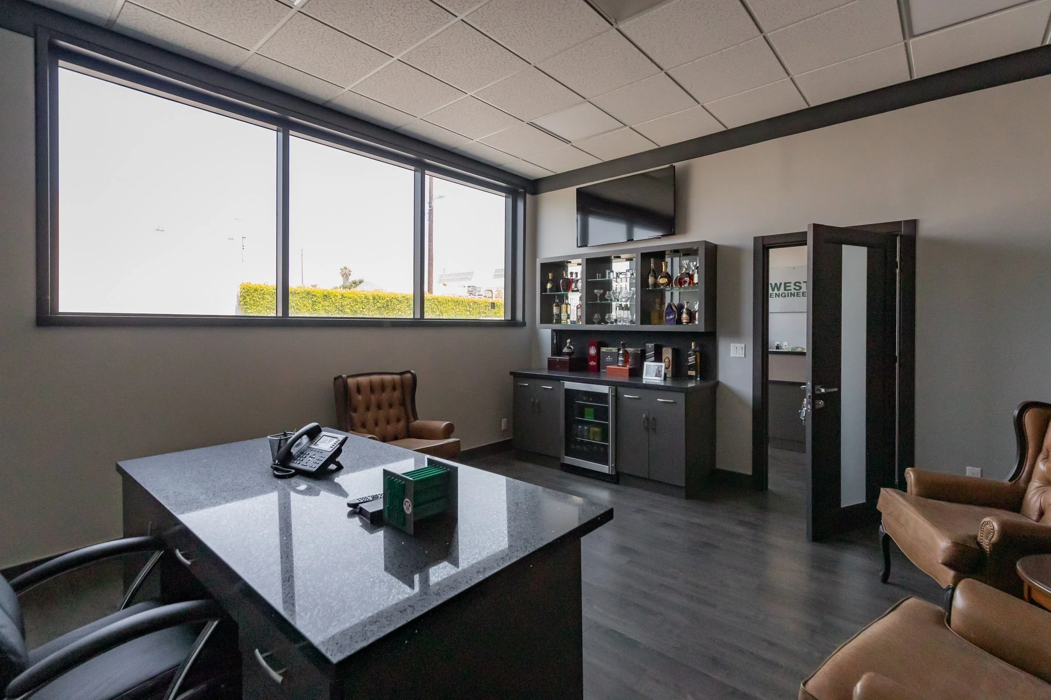 Office space with large window, brown leather chairs, a black countertop table with a telephone, a cabinet with various bottles and glasses, and a flat-screen TV on the wall.