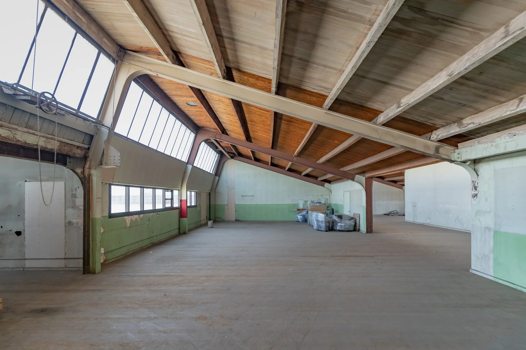 Empty, unfinished indoor space with wooden floors, large windows, exposed beams, and partially painted walls.
