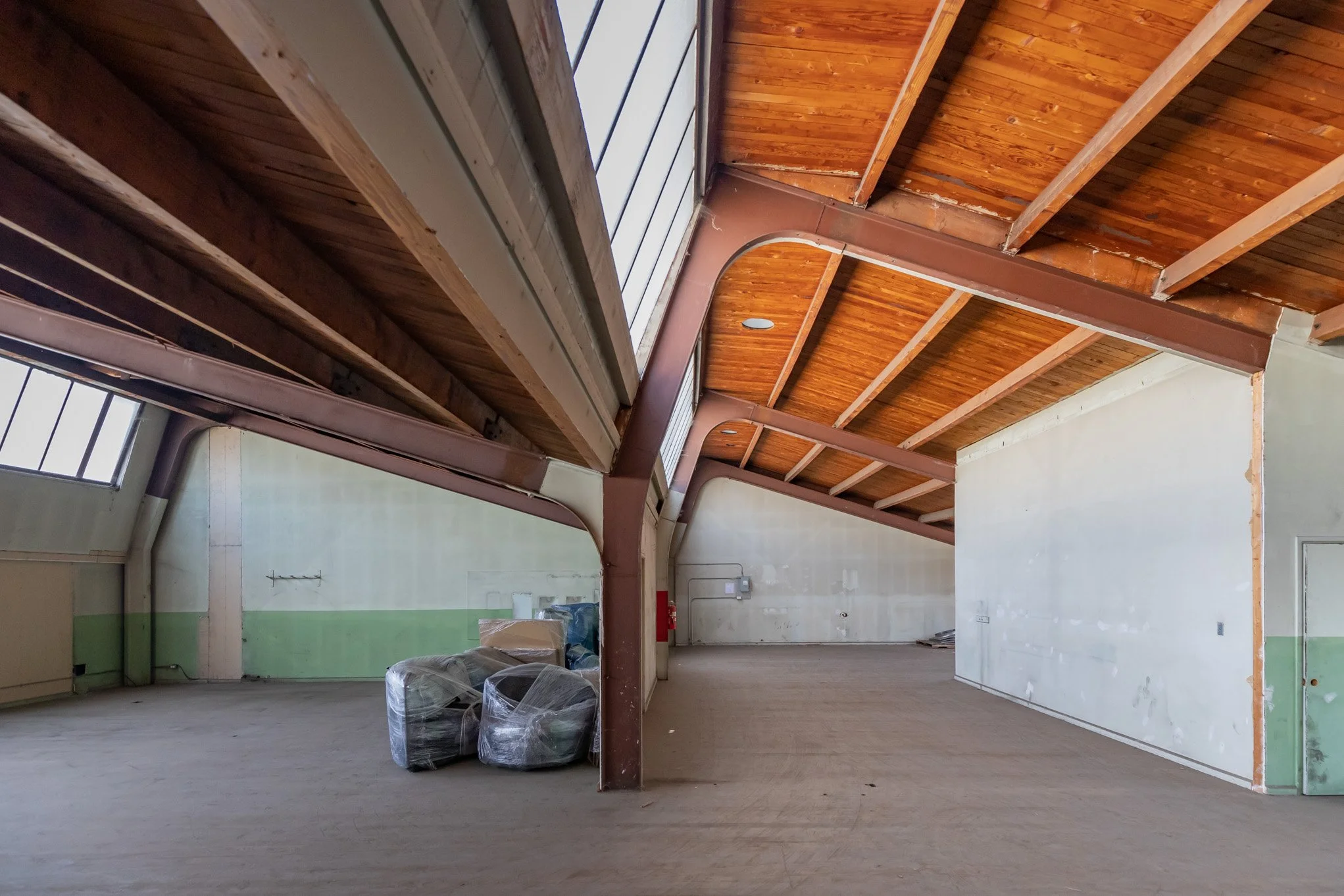 Empty interior space with exposed wooden ceiling, large windows, and some wrapped items in the corner.