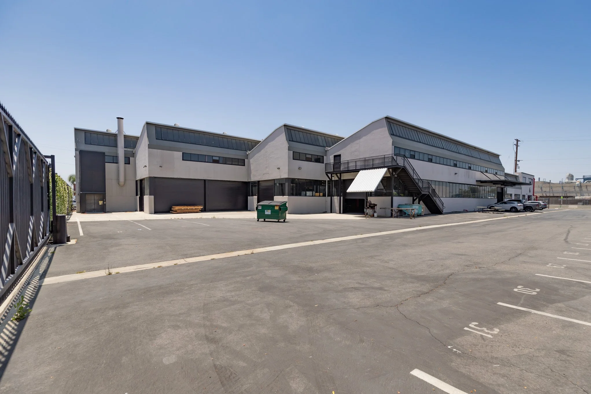 An empty parking lot in front of a modern, multi-story building with large windows and a staircase leading to an upper level, under a clear blue sky.