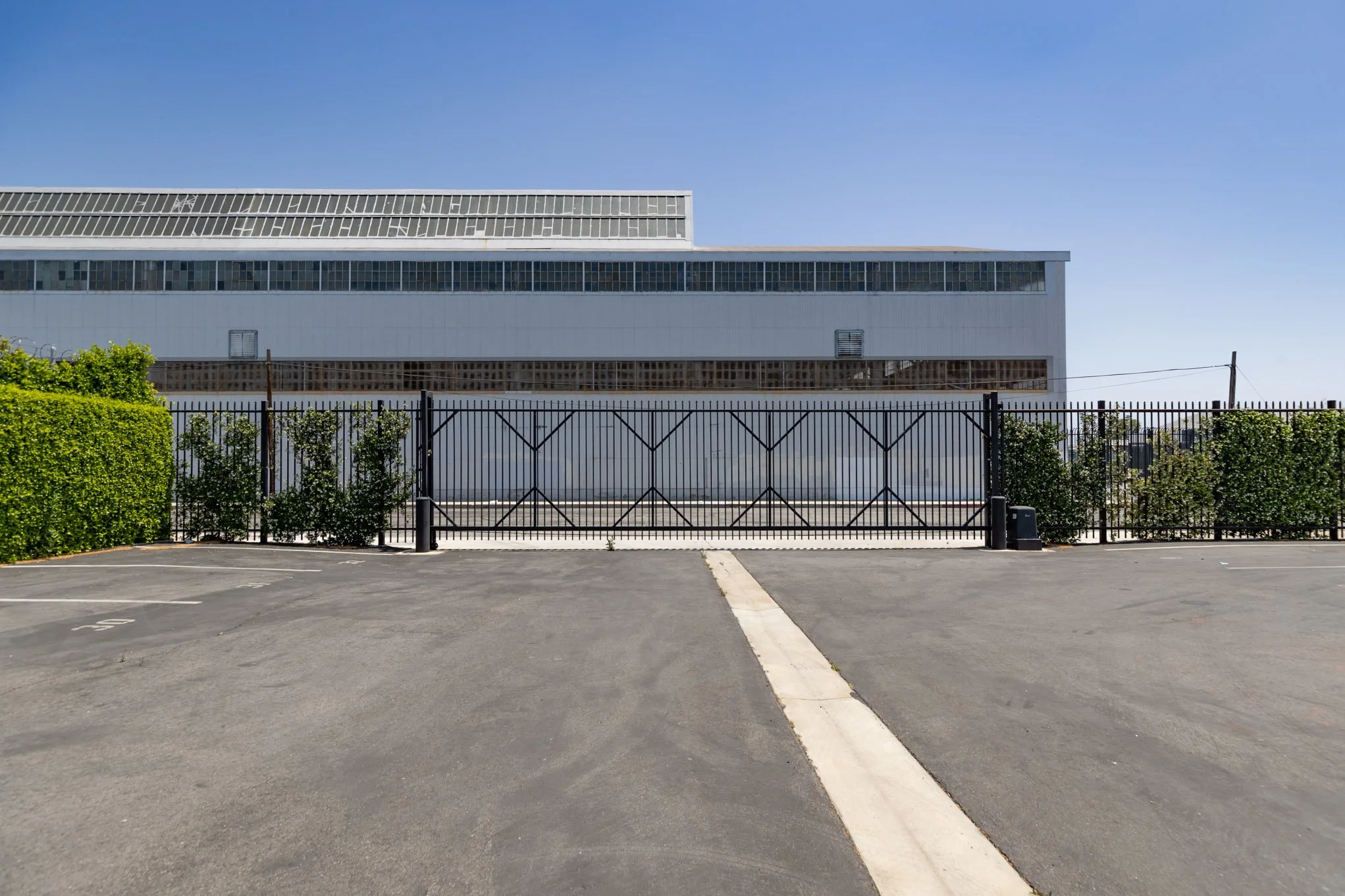 Empty parking lot with a gated fence in front of a large industrial or warehouse building under a clear blue sky.