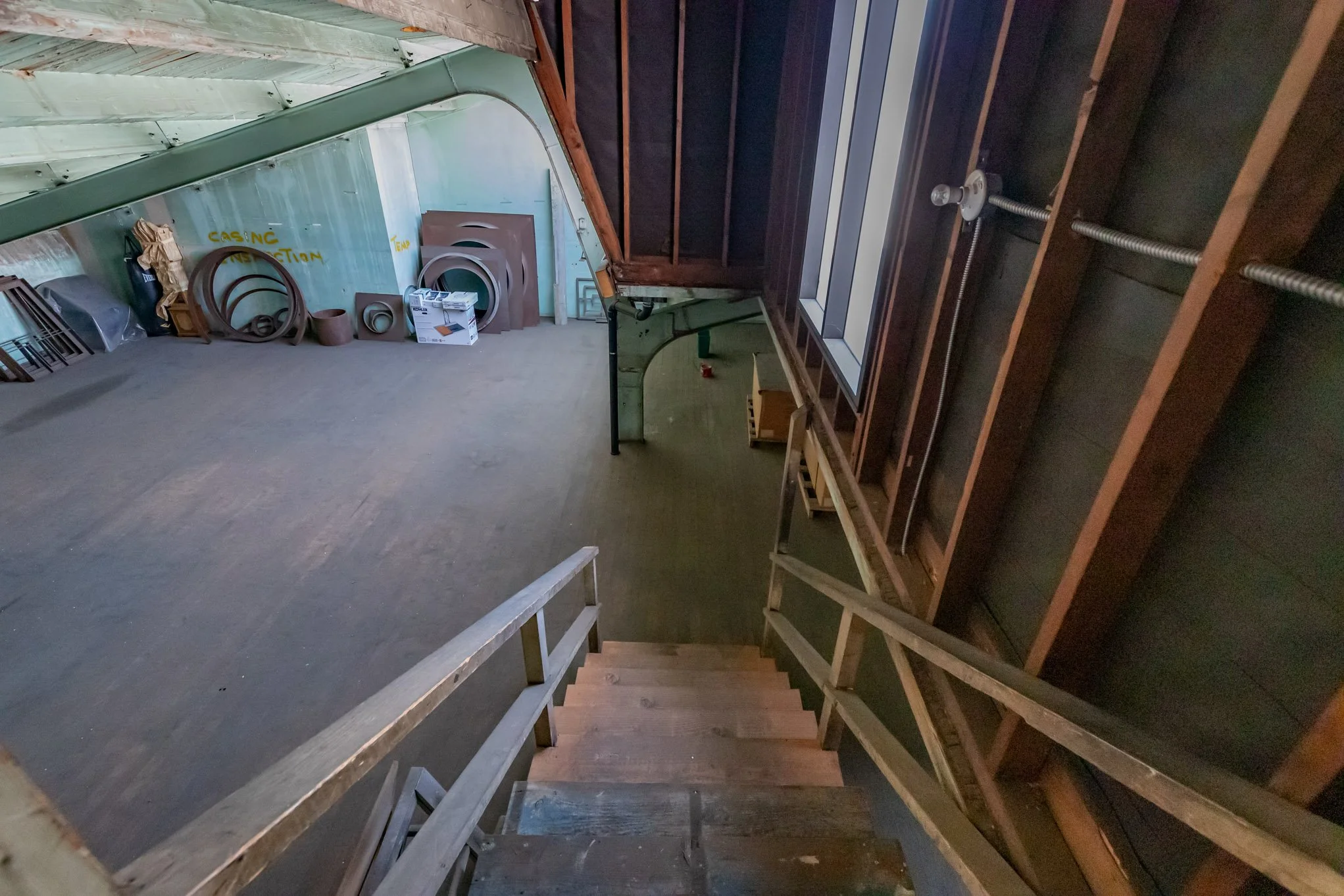 View of an attic space from the top of a staircase, showing wooden stairs and unfinished walls with exposed beams and insulation, along with storage items such as round frames and boxes.