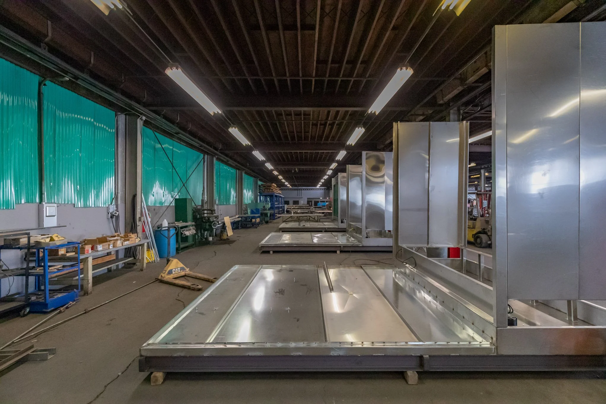 Industrial warehouse with metal structures, workbenches, and overhead lighting, with green fabric coverings on the left side and equipment in the background.