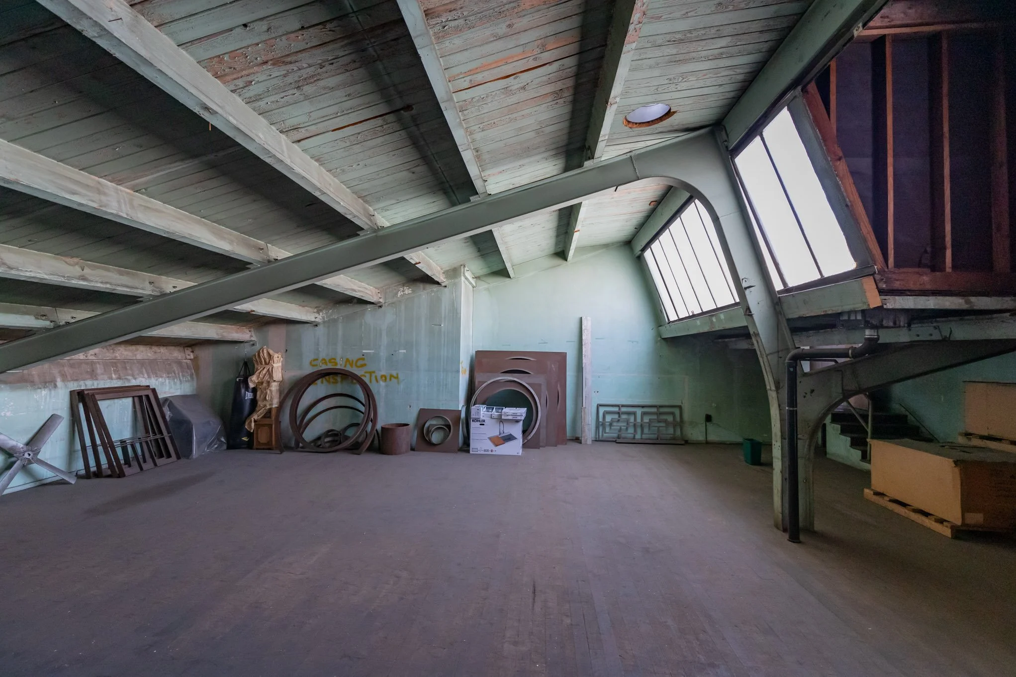 Empty attic room with wooden floor, green walls, and sloped ceiling with exposed beams, containing various household items and furniture stored along the walls.