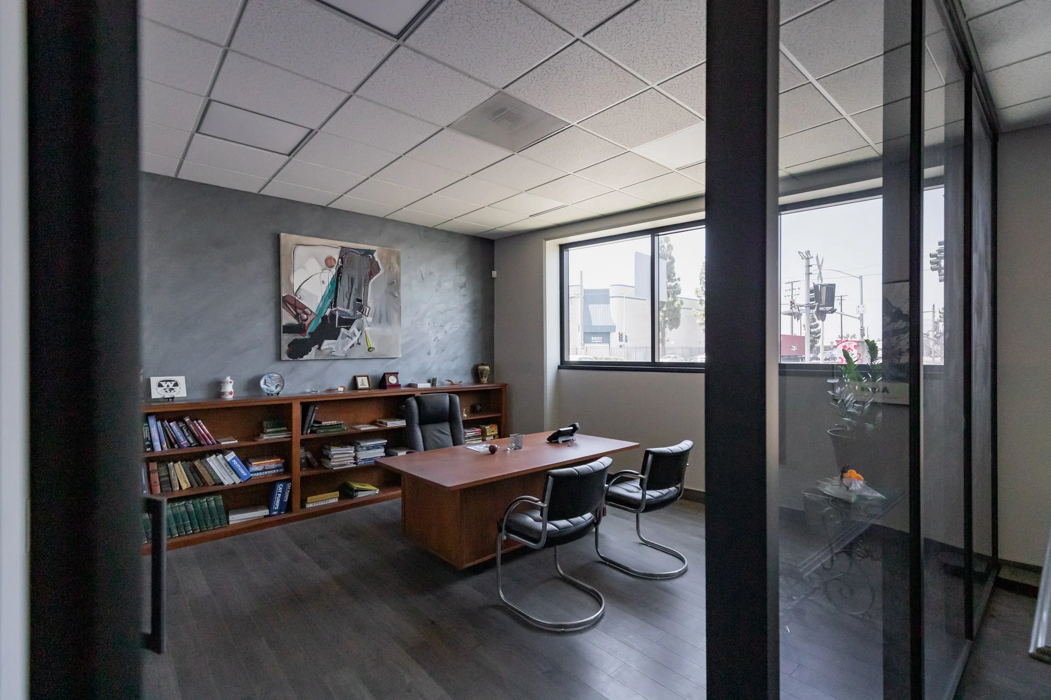 Empty office with a wooden desk, three chairs, a bookshelf filled with books and decorative items, a large window, and a wall-mounted abstract painting.