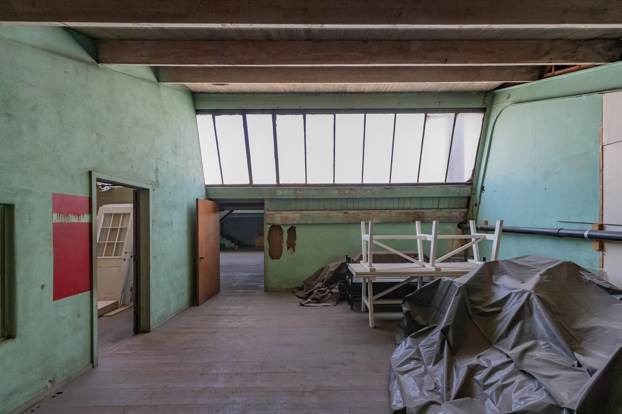 An empty room with green walls, wooden flooring, and a slanted ceiling with large skylights. There are some stacked white tables or furniture, covered items, and a door that is partially open, revealing another room with a staircase.