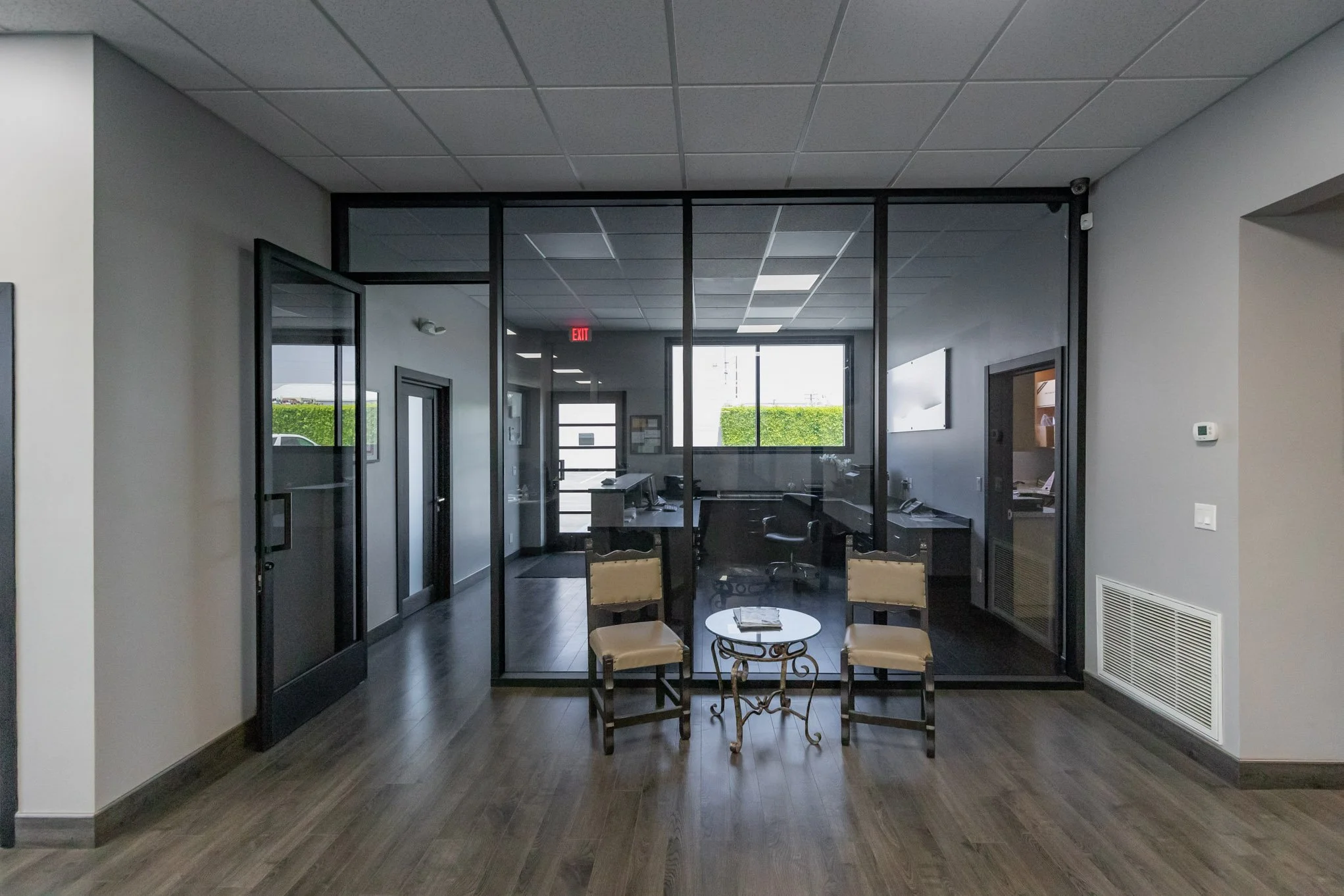 Office reception area with three chairs and a small round table, separated from main office by glass walls, with desks, chairs, and large windows visible inside.