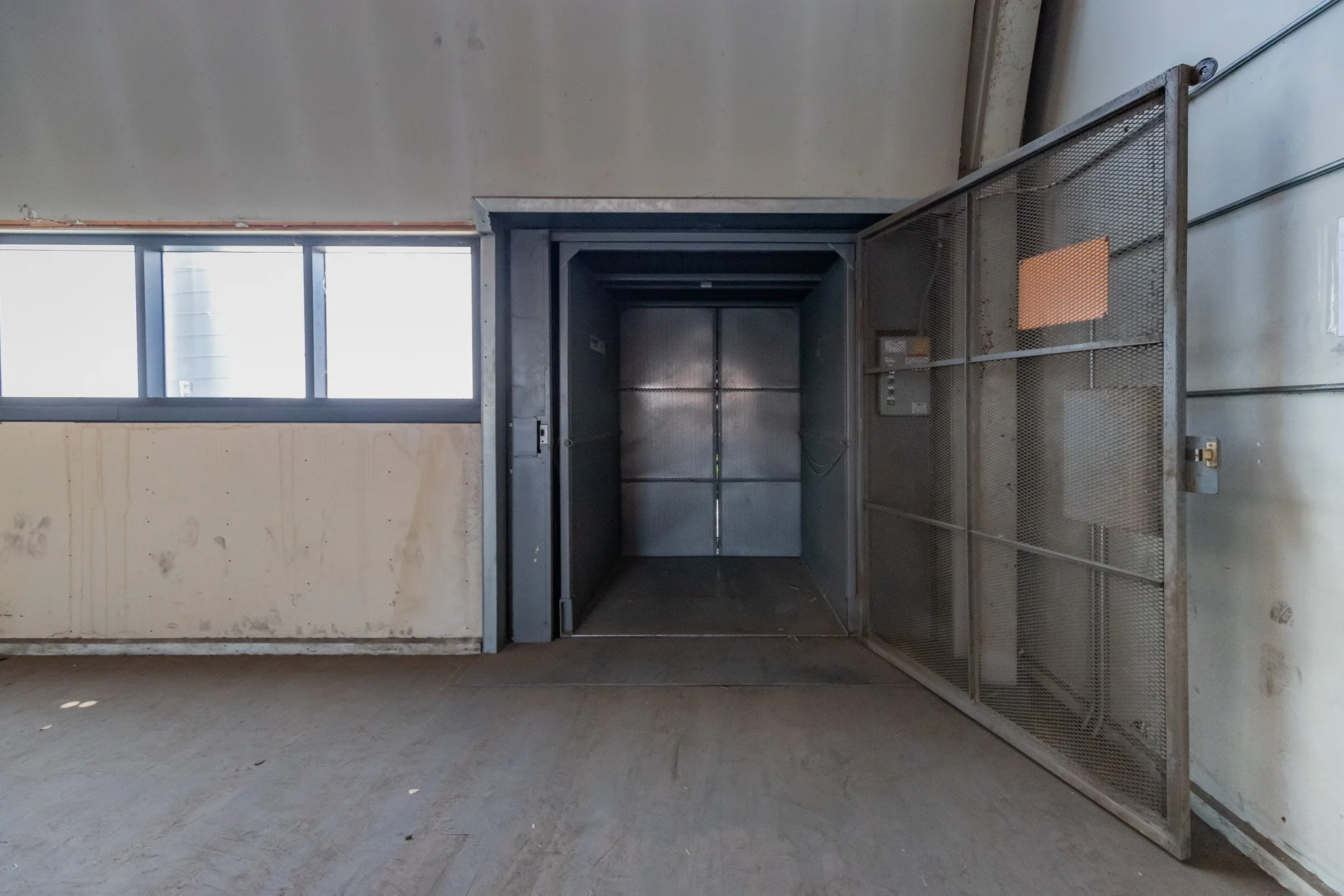 An empty industrial elevator with metal interior, open gate, near a window in a worn building.