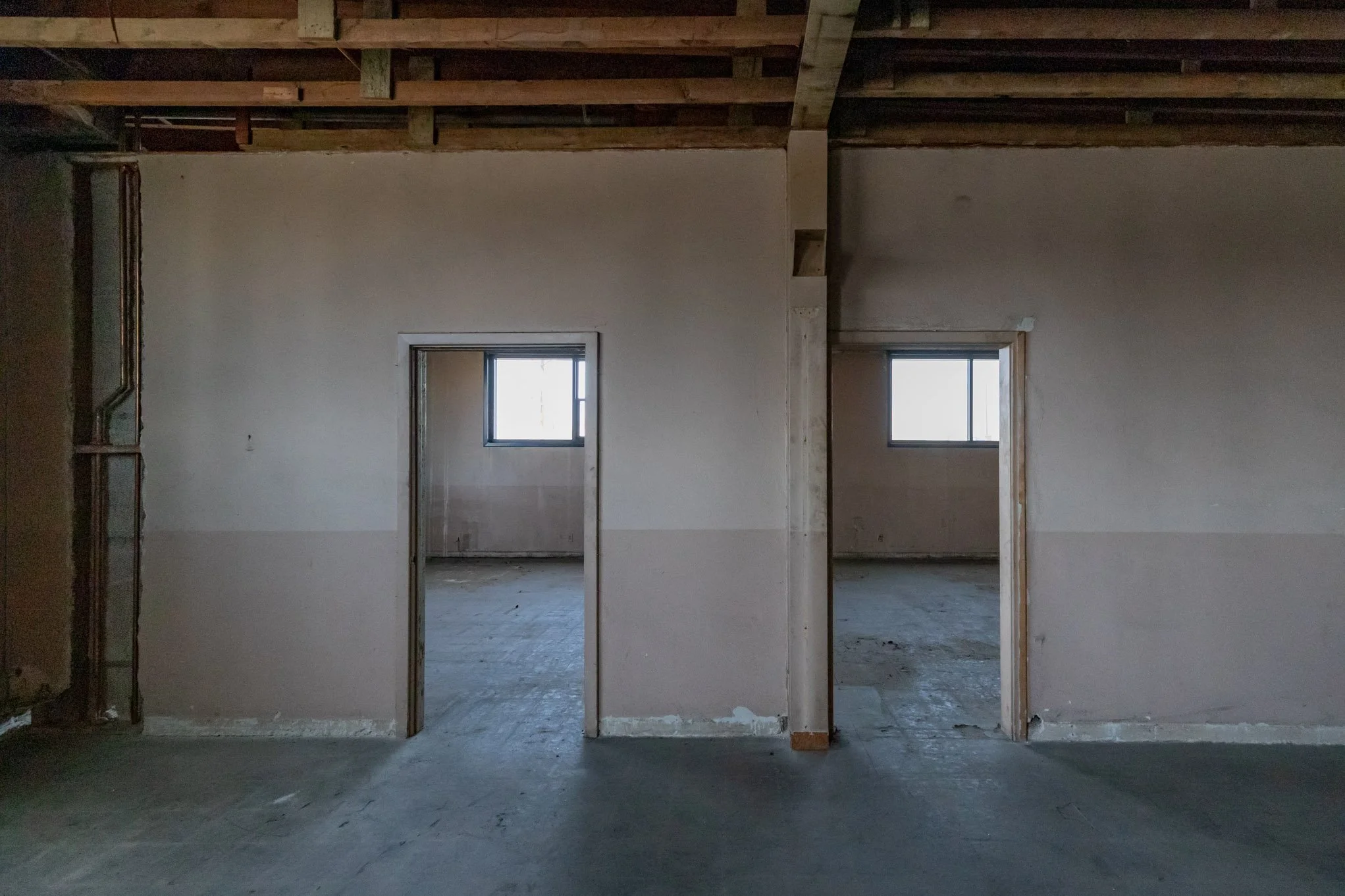 Interior view of a building under construction with two doorways and two windows, exposed ceiling joists, and unfinished walls.