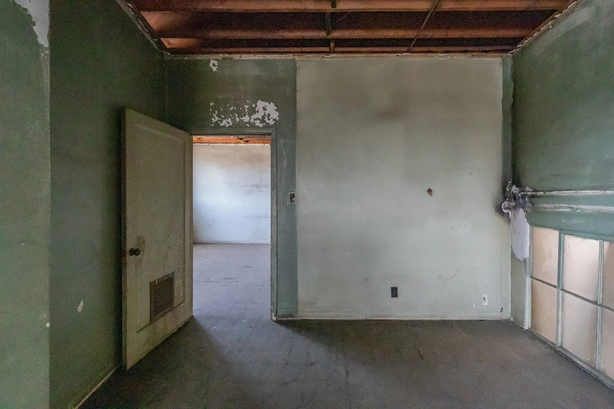 Interior of an abandoned room with peeling green paint, a partially open door, and damaged ceiling and walls.