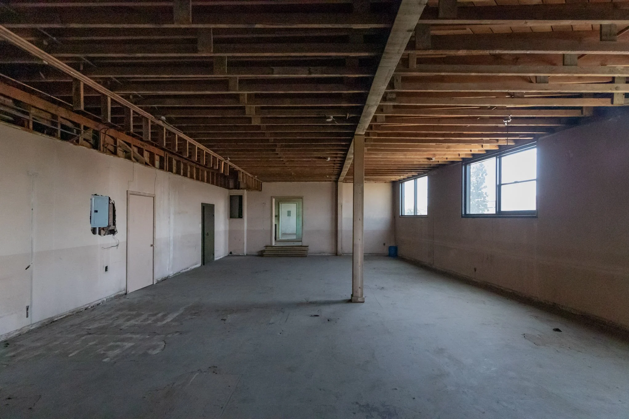 An unfinished interior space with exposed wooden ceiling beams, large windows, and bare concrete floors. Some electrical wiring and boxes are visible on the wall.