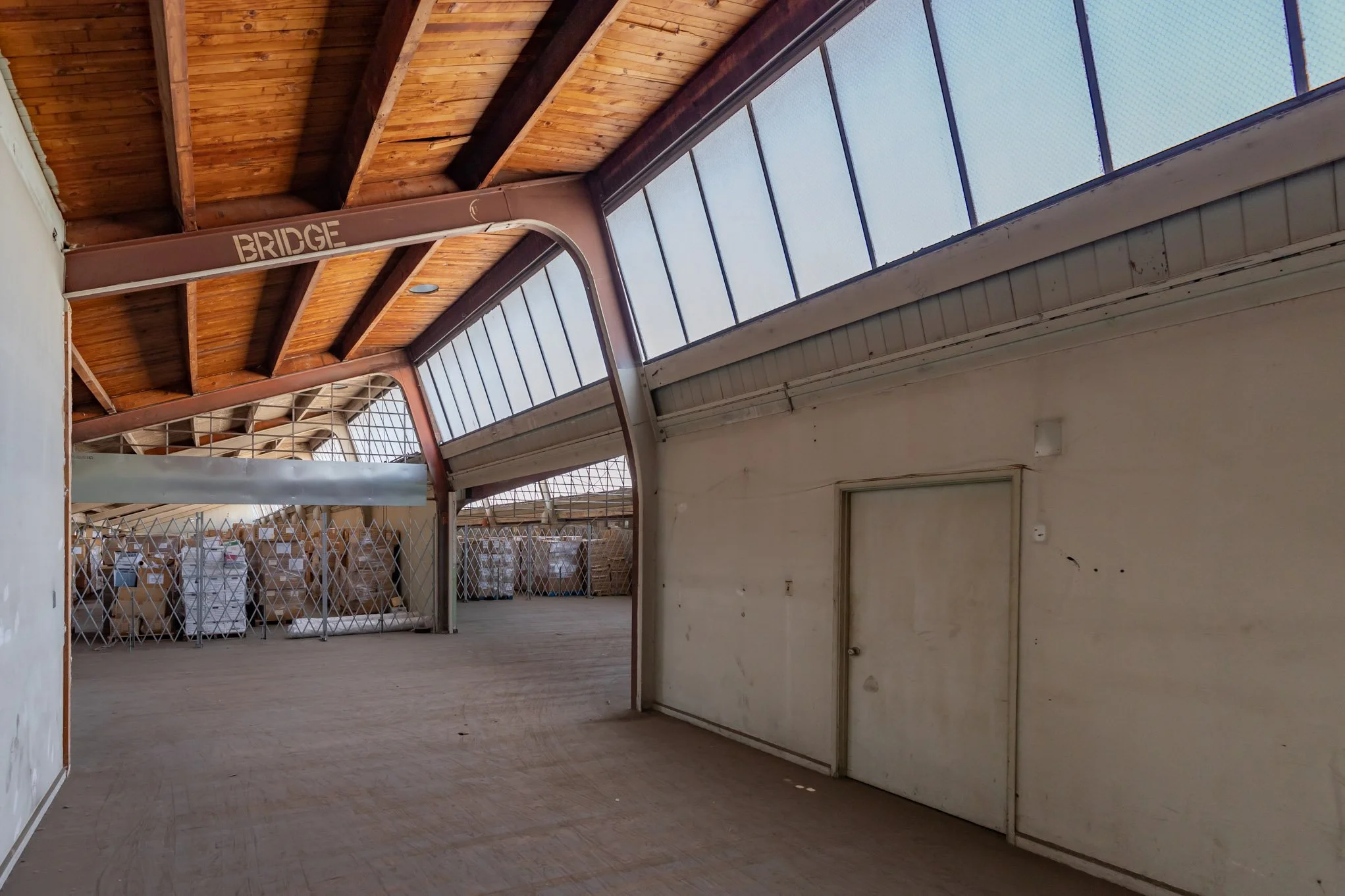 Indoor warehouse space with high wooden ceiling, metal beams, and a large window wall, with some stacked boxes or pallets visible in the background.