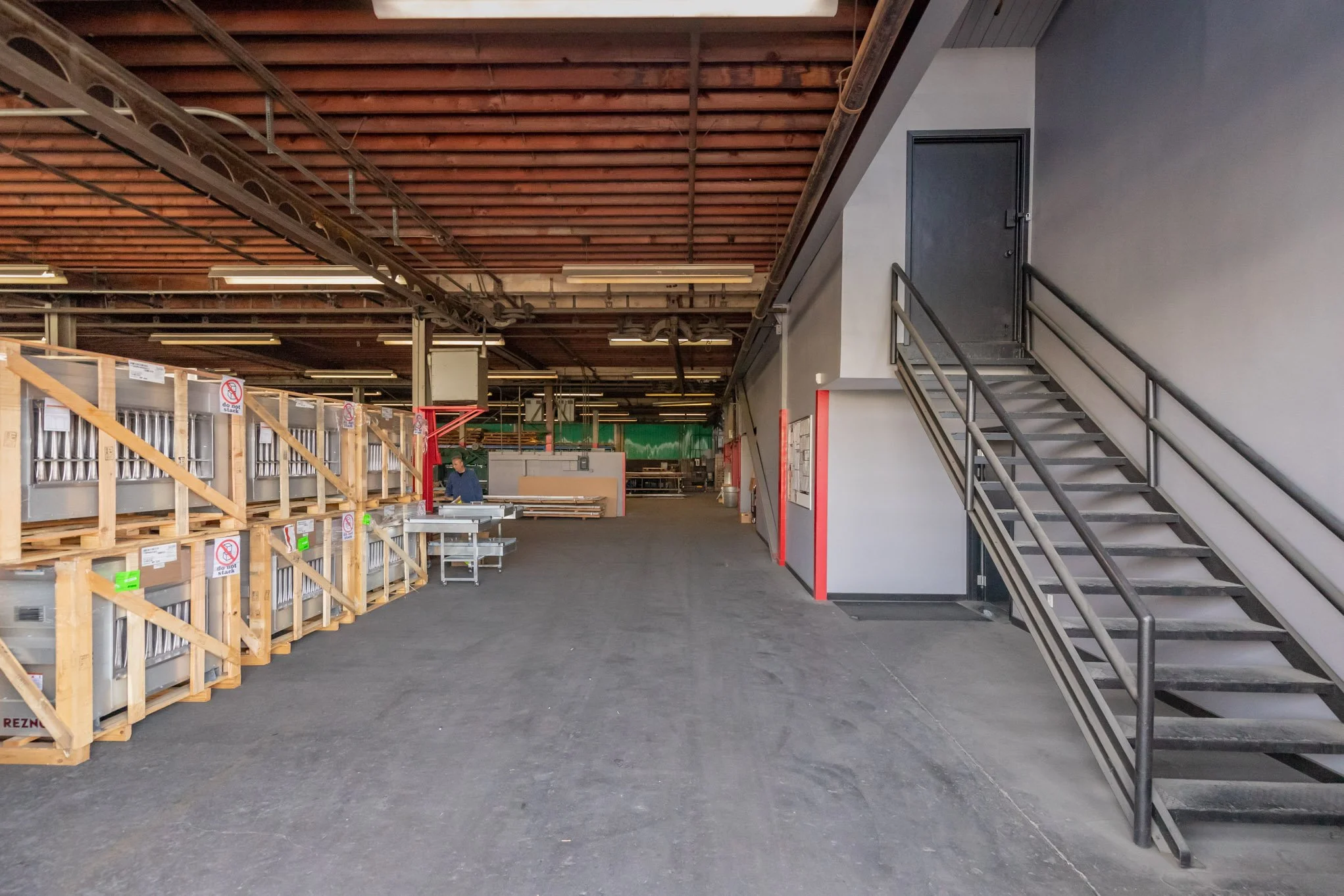 Interior of a warehouse or industrial space with stacked items on the left, a staircase with metal railings on the right, and a person working near a metal table in the background.
