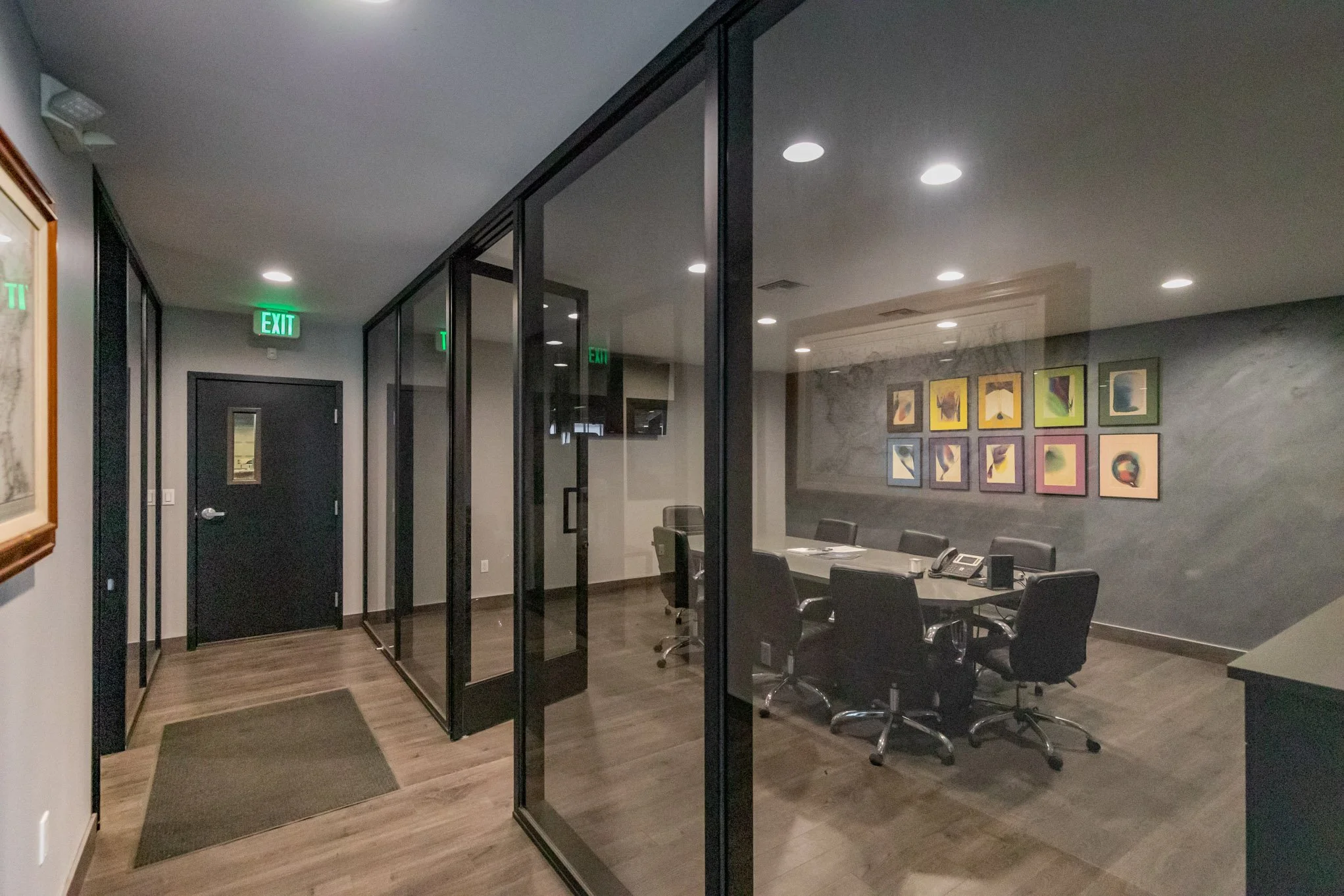 Empty conference room with a large table surrounded by black chairs, framed colorful abstract art on the wall, conference phones, and a glass wall and door.