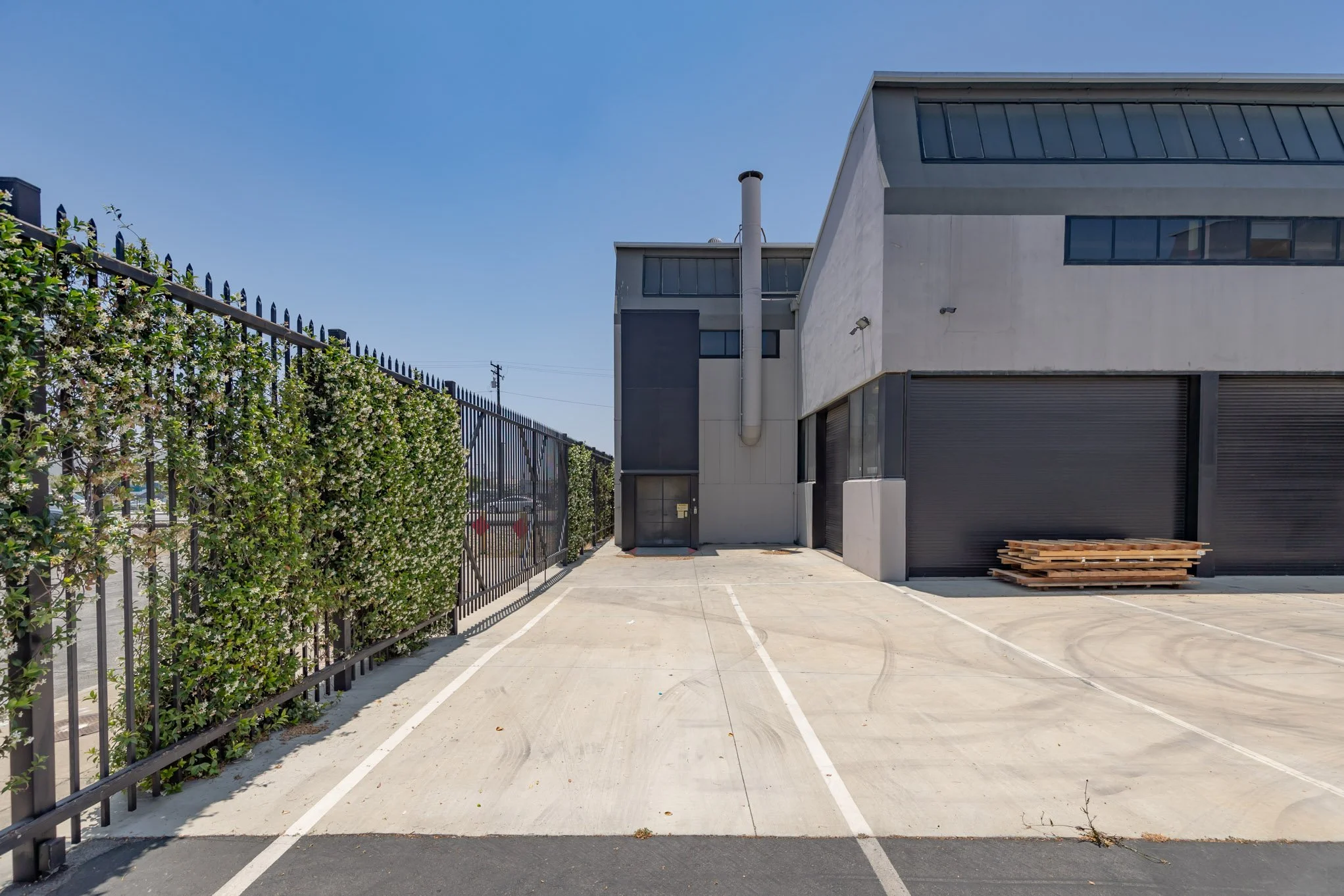 Empty parking lot with three parking spaces, a building with black roll-up doors, a stack of wooden pallets, a black gate, and a hedge on the left side under a clear blue sky.