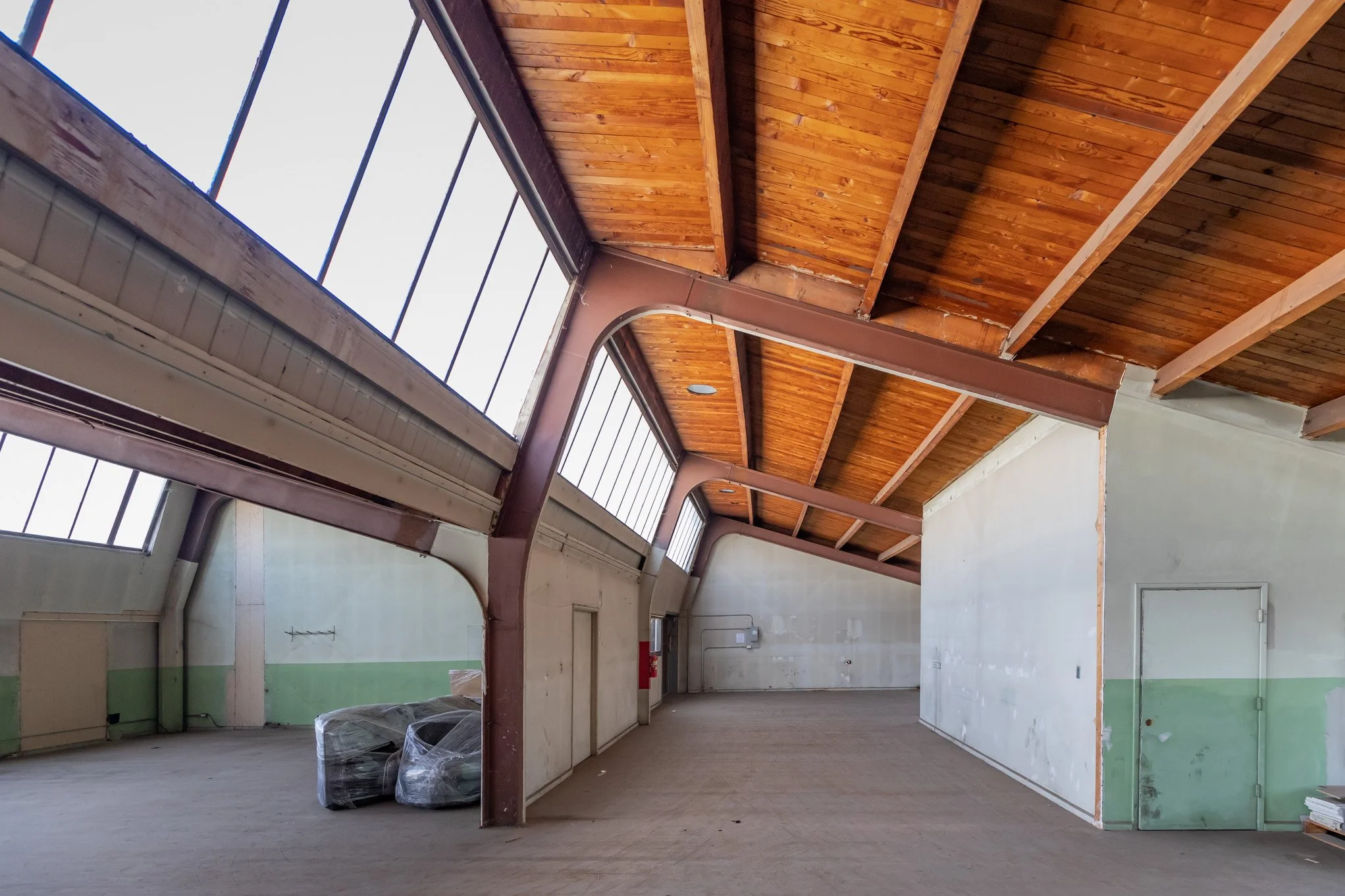 Empty industrial space with a high wooden ceiling, large windows, and some wrapped objects on the floor.