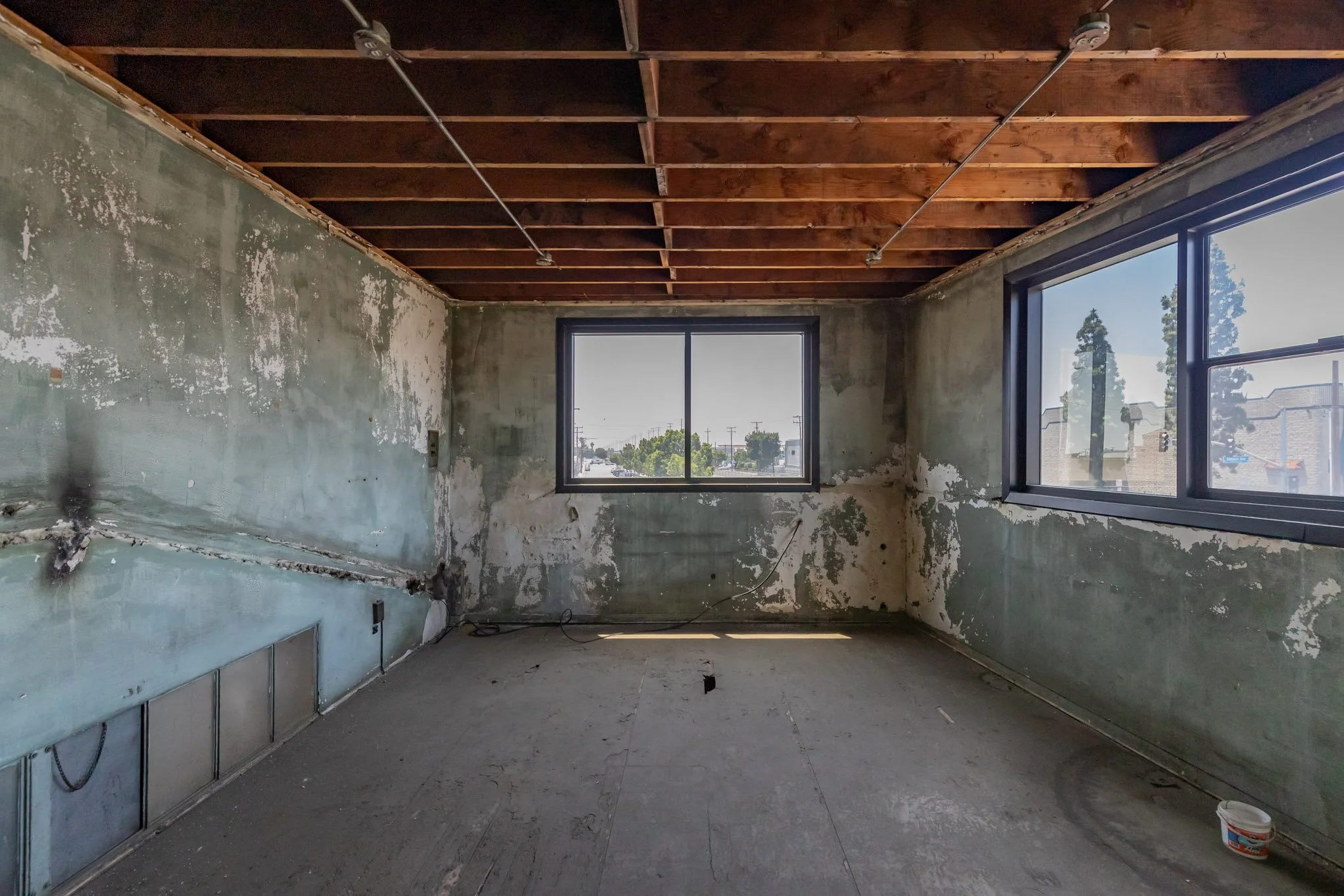 Interior of a room under renovation with exposed wooden ceiling beams, unfinished walls with peeling paint, and large windows showing an outdoor street scene.
