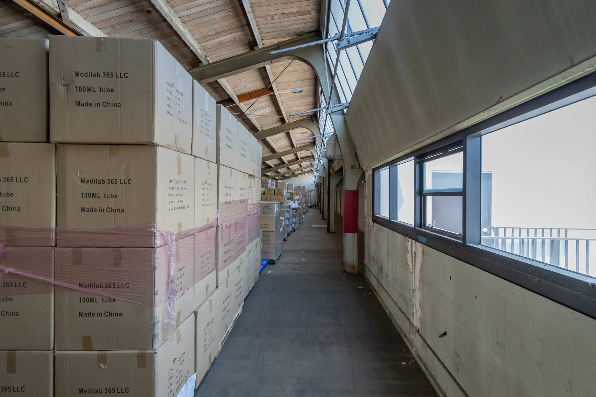 Corridor with boxes stacked along the wall inside a building, with a row of large windows on the right side.